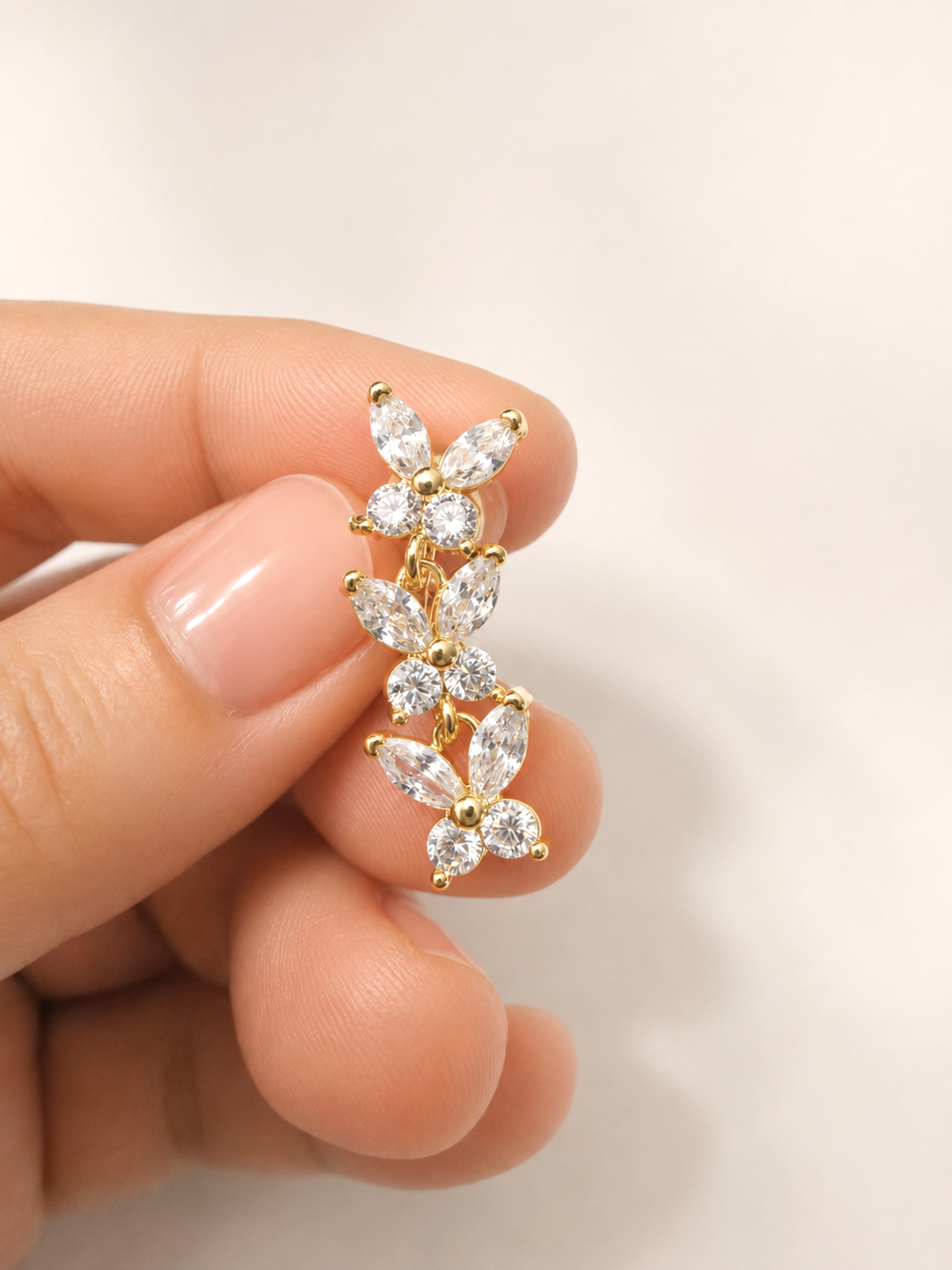 A close-up of a hand holding a small gold floral stud earring with clear marquise-cut gemstones against a soft off-white background.
