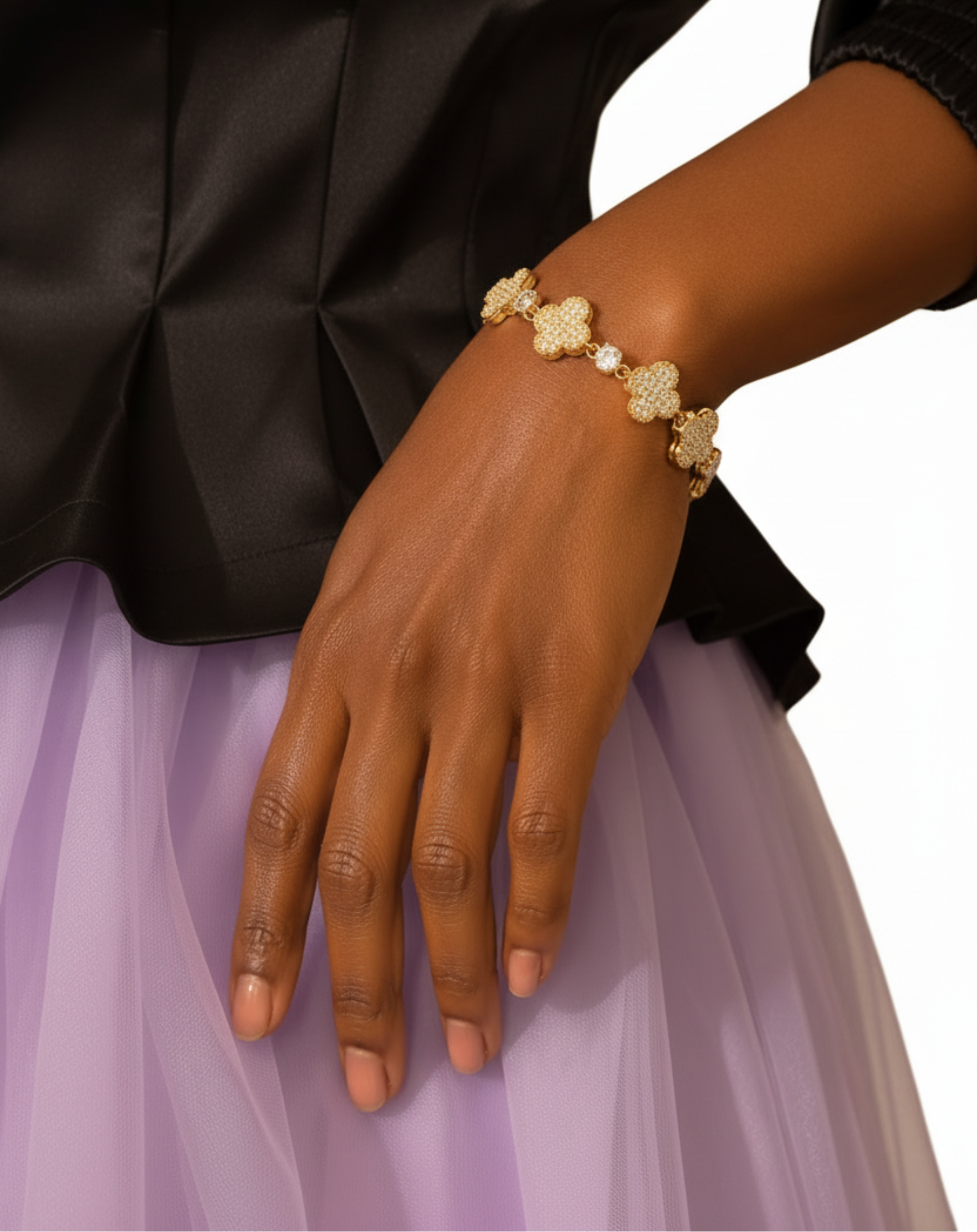 A close-up of a person's wrist wearing a gold bracelet with four clover-shaped links, each adorned with small clear stones. The person is wearing a black satin blouse and a lavender skirt, with a woven cane chair visible in the background.