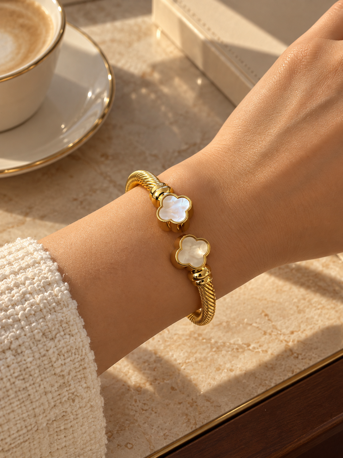A gold-toned cuff bracelet with two white mother-of-pearl flower charms, shown worn on a person's wrist in a warm, sunlit cafe setting with a coffee cup in the background.