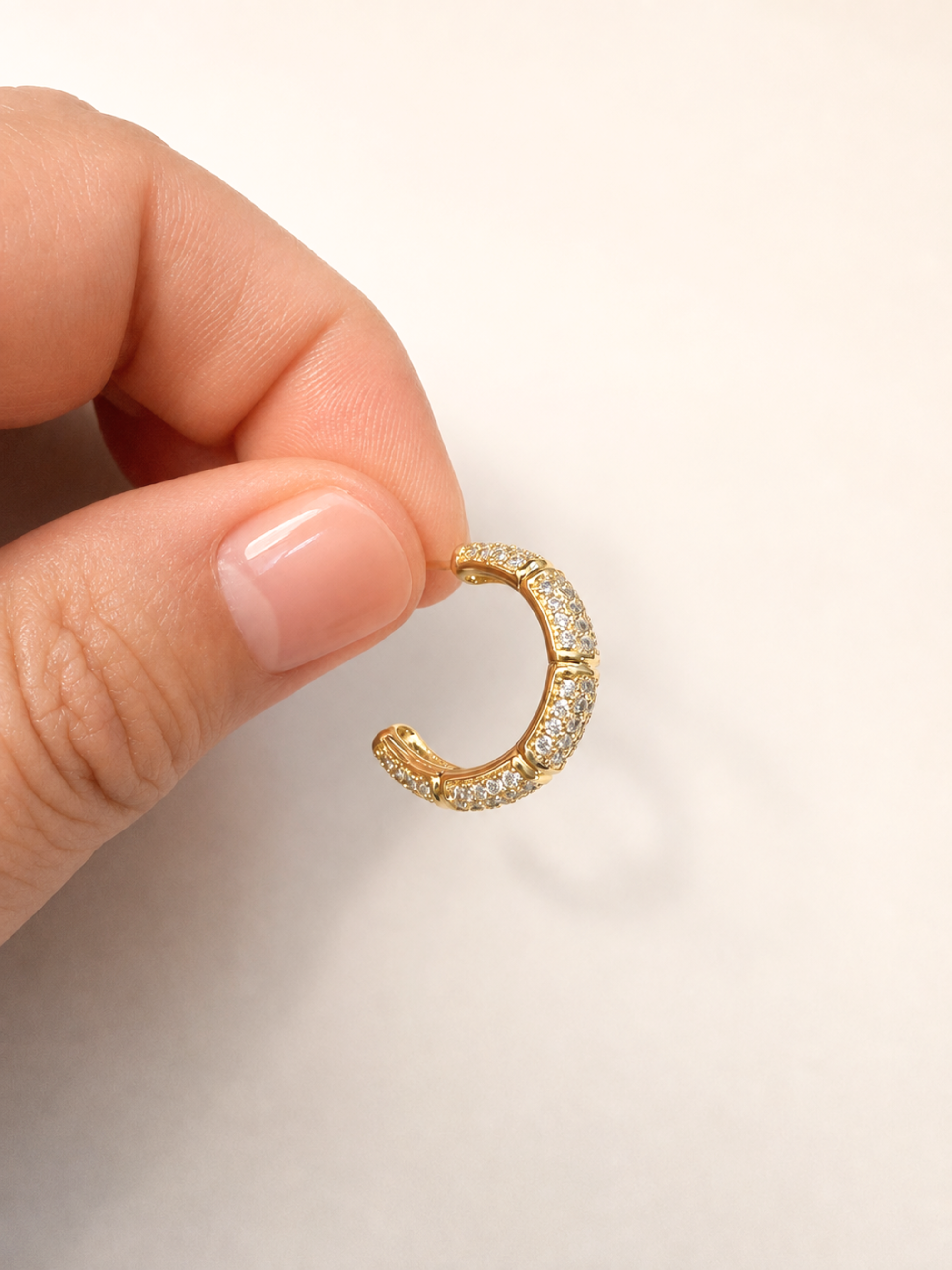 A close-up of a hand holding a small gold huggie hoop earring paved with clear stones against a plain light background.