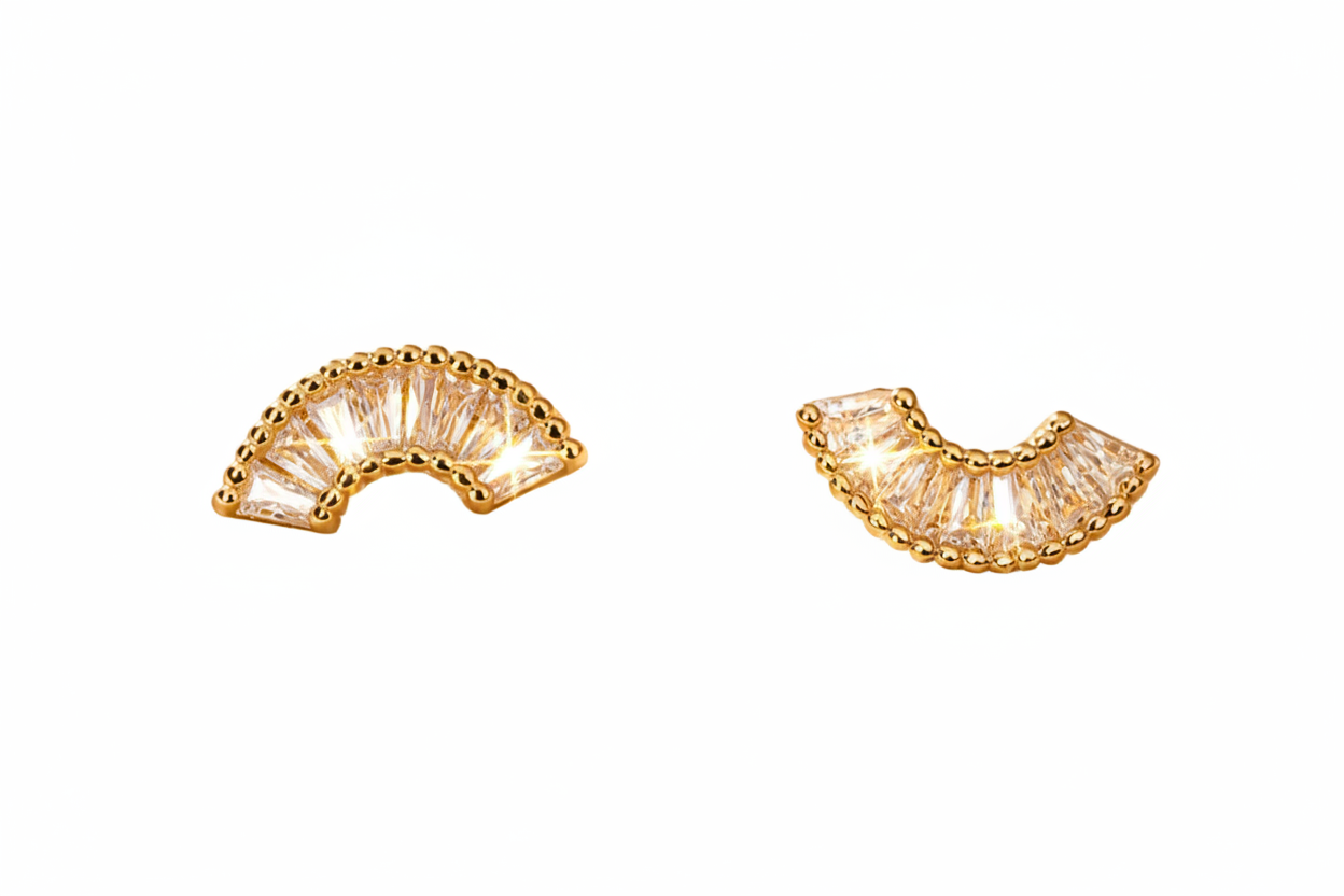 A pair of gold crescent-shaped stud earrings with clear baguette crystals, displayed on a draped champagne-colored satin fabric.
