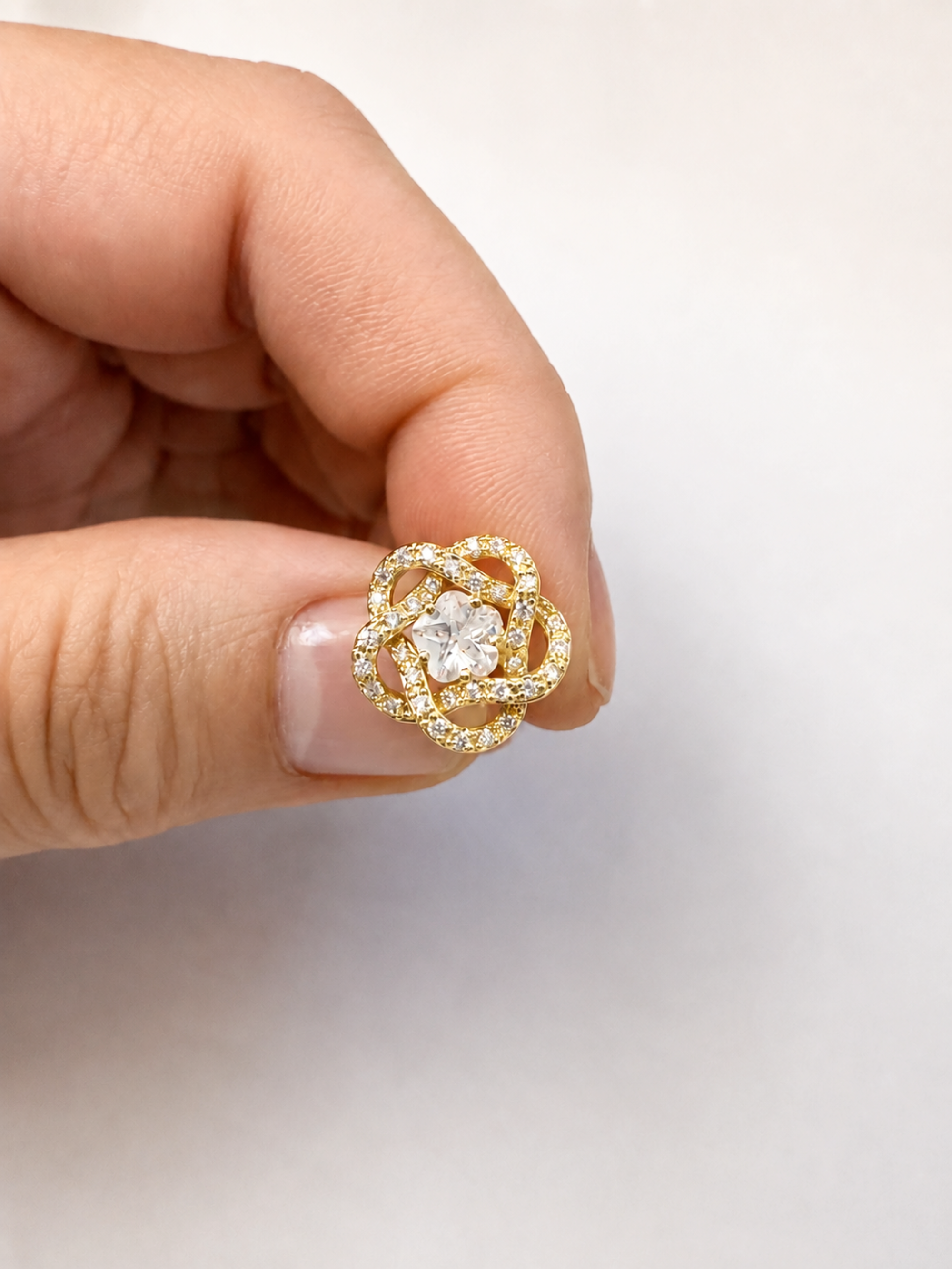 A hand holding a small gold flower-shaped ring with a clear center stone and surrounding pave-set stones, shown against a plain light background.