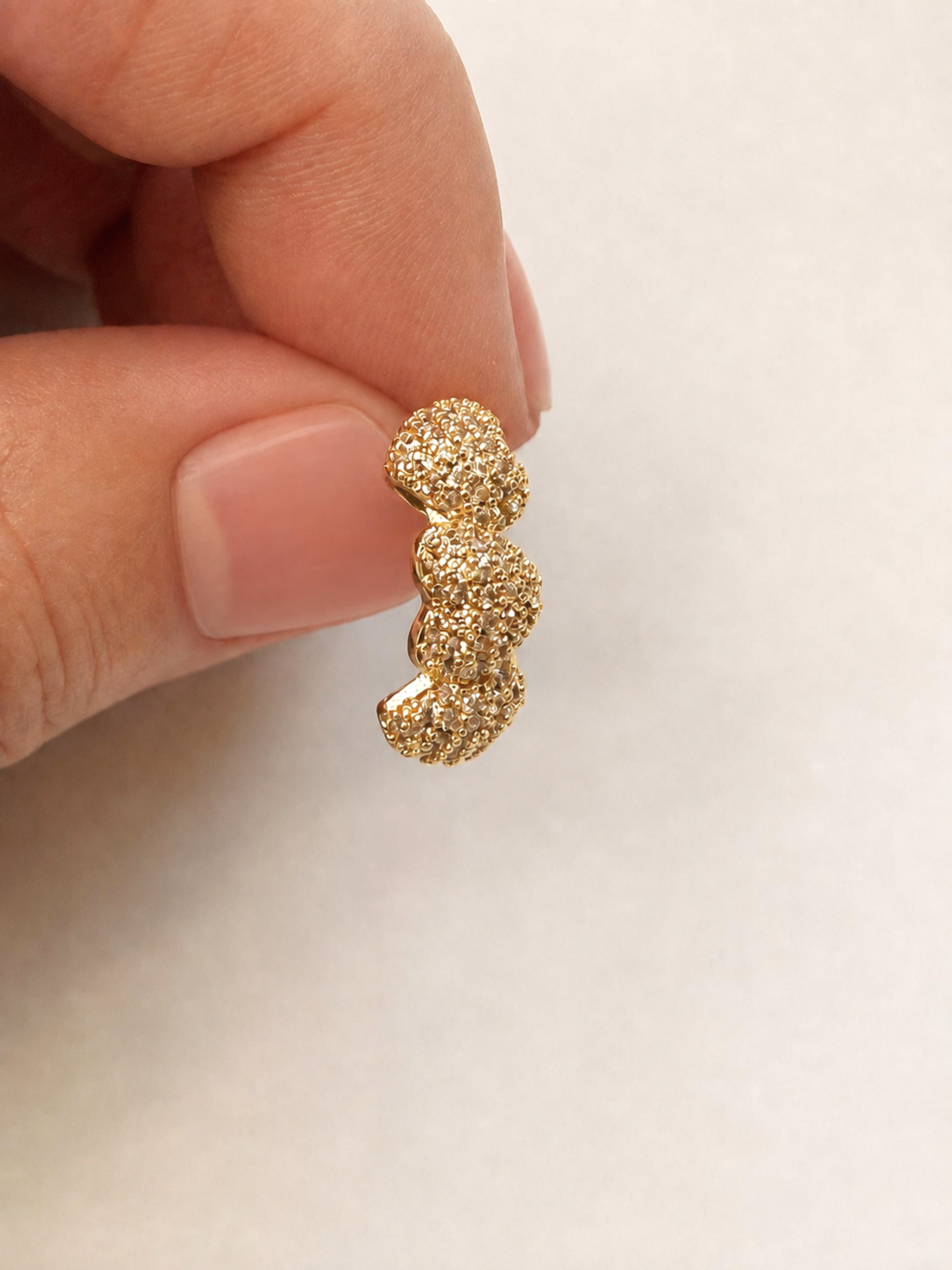 A close-up of a hand holding a small gold textured earring against a soft light gray background.