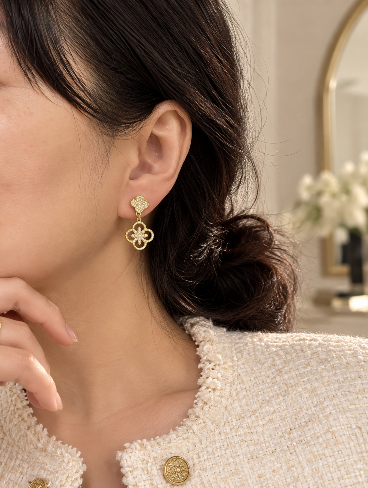 A close-up of a woman wearing a delicate gold dangle earring with a small floral top and a four-loop charm. She is wearing a textured cream tweed jacket, and the background shows a softly blurred indoor setting with a mirror and flowers.
