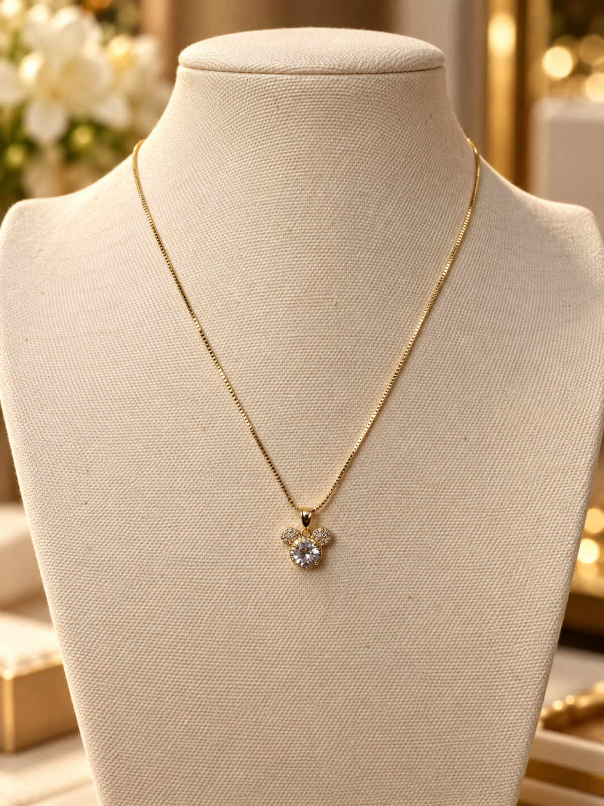 A gold chain necklace with a Mickey Mouse-shaped pendant set with a clear central gemstone, displayed on a beige fabric jewelry bust.