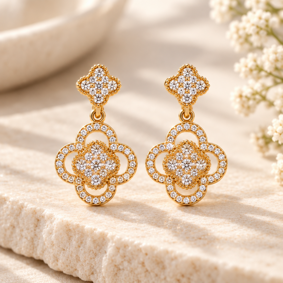 A pair of gold-toned drop earrings with intricate pavé crystal clover motifs, displayed on a shimmering beige fabric surface with soft-focus decorative elements in the background.