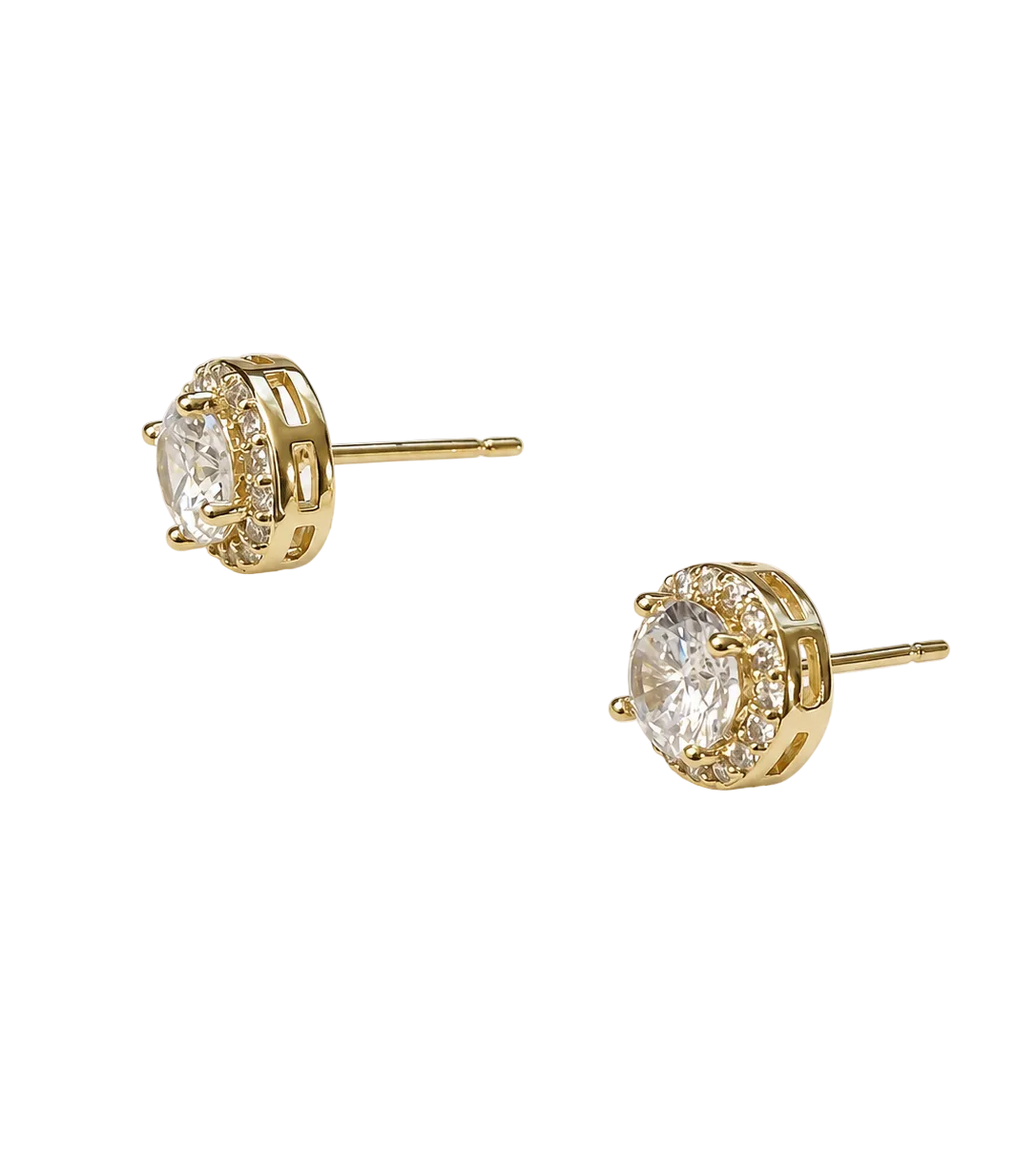 A pair of gold stud earrings with round clear gemstones in a halo setting, shown on a plain off-white background.