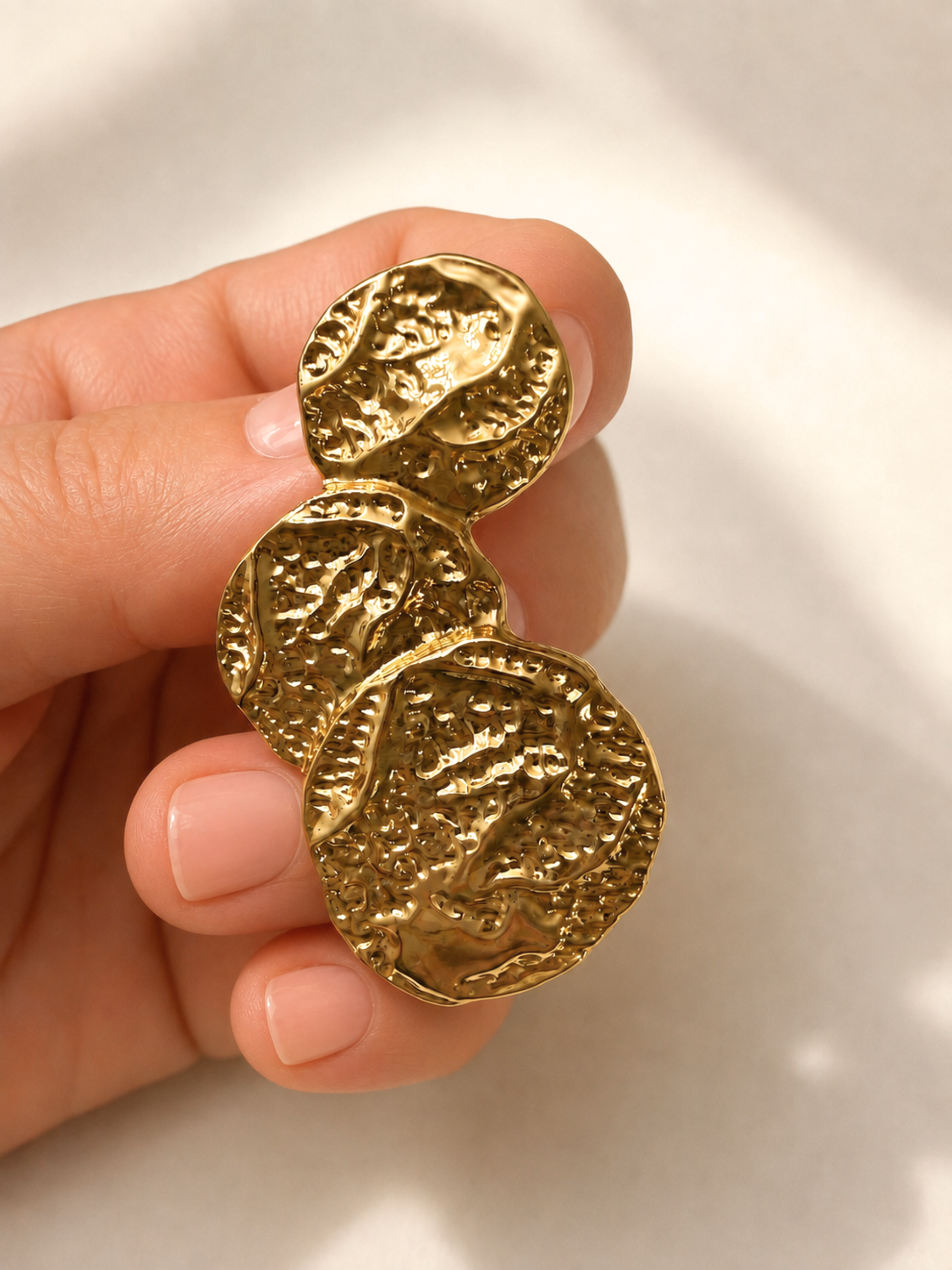A close-up of a hand holding a gold-toned jewelry piece with three stacked, textured coin-like discs against a soft light background.