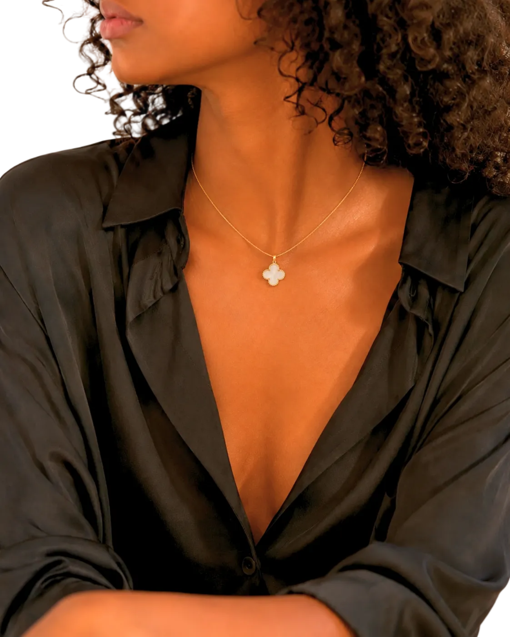 A close-up of a model wearing a delicate gold chain necklace with a small white clover-shaped pendant, styled with a black satin shirt.
