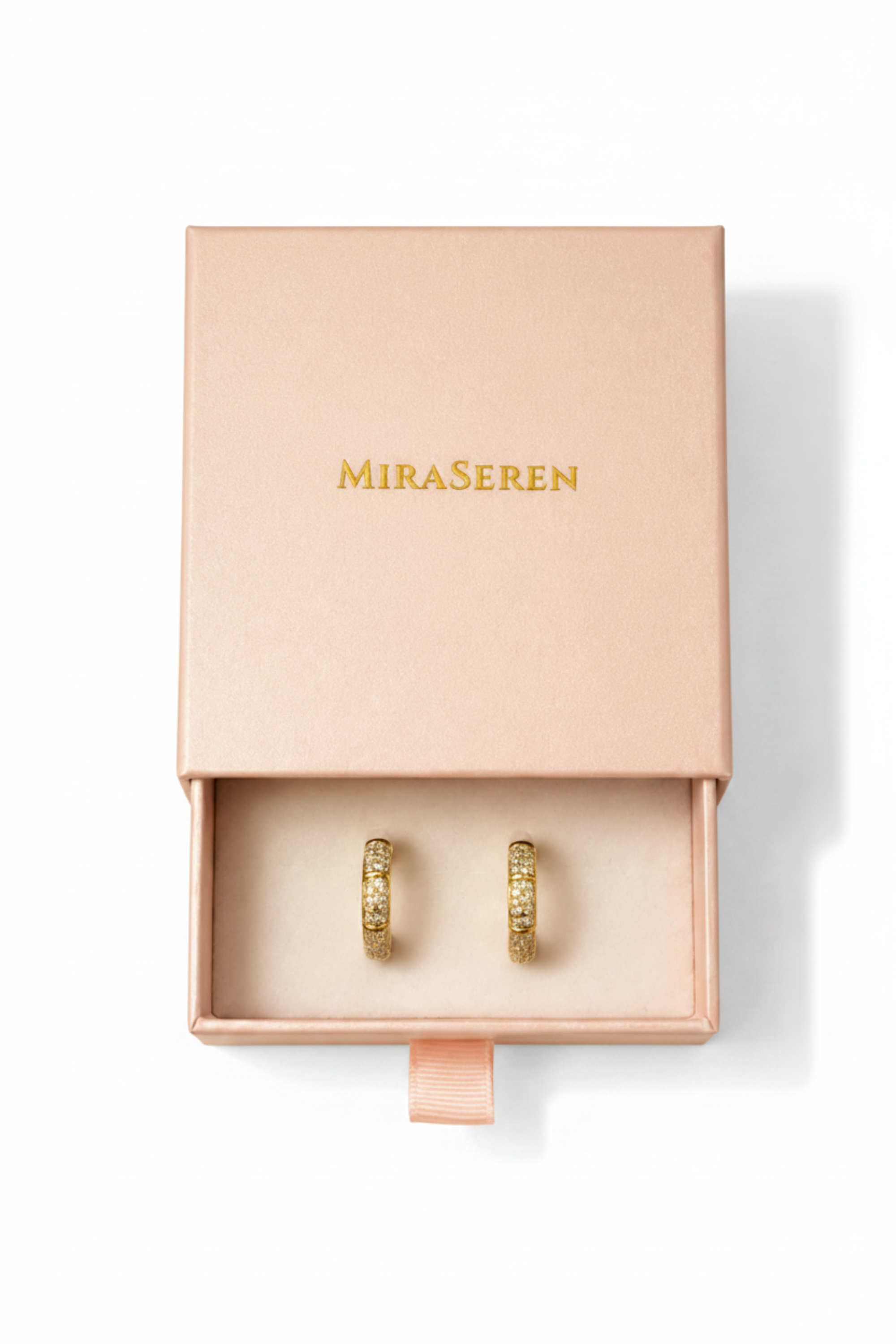 A pair of gold rings displayed in an open blush pink Miraseren jewelry box, with the matching pink lid resting above on a warm beige background.