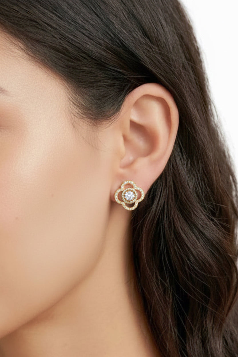 A close-up of a model's ear wearing a gold stud earring with a central clear gemstone surrounded by small sparkling stones.