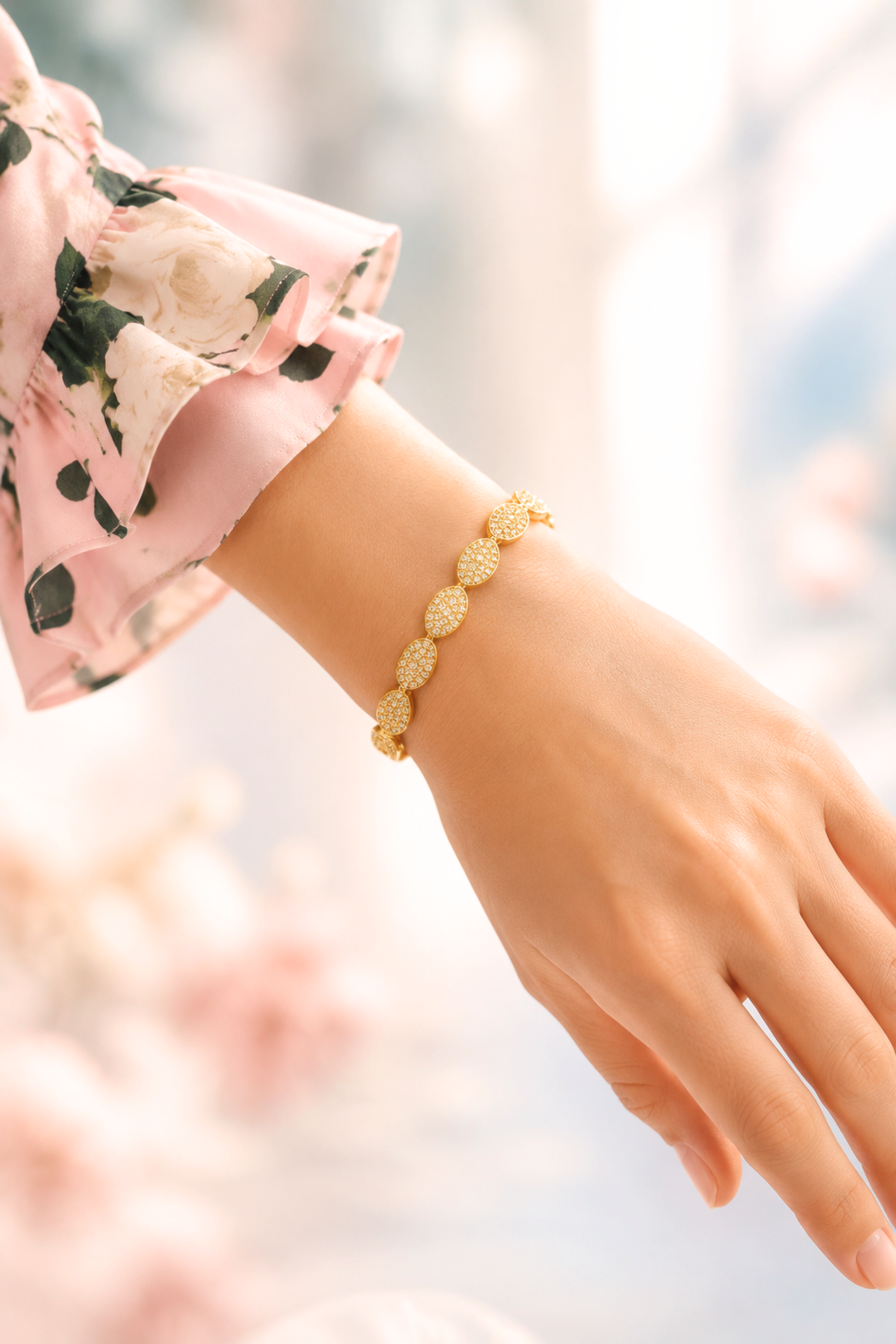 A gold link bracelet with textured oval links worn on a woman's wrist, shown in a soft-focus lifestyle setting with a pink floral sleeve visible.
