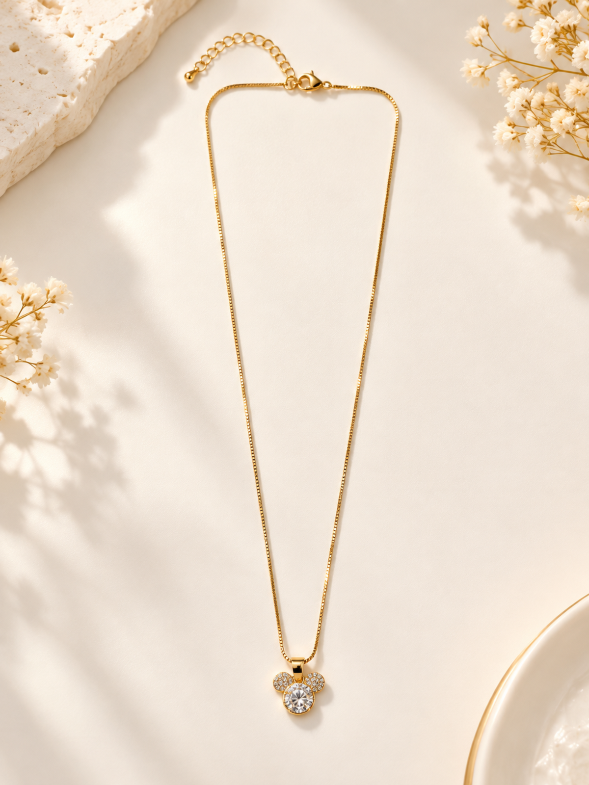 A gold chain necklace with a Mickey Mouse-shaped pendant set with clear crystals, displayed on a warm cream surface with decorative dried flowers and stone props.