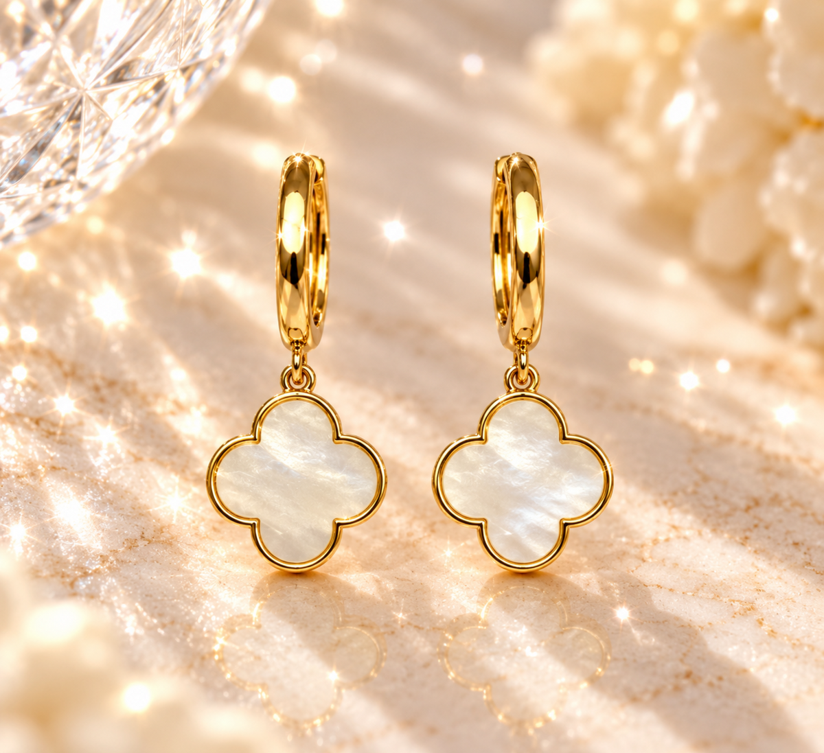 A pair of gold hoop earrings with four-leaf clover-shaped dangles made from iridescent mother-of-pearl, displayed on a shimmering gold marble surface with a crystal ball in the background.