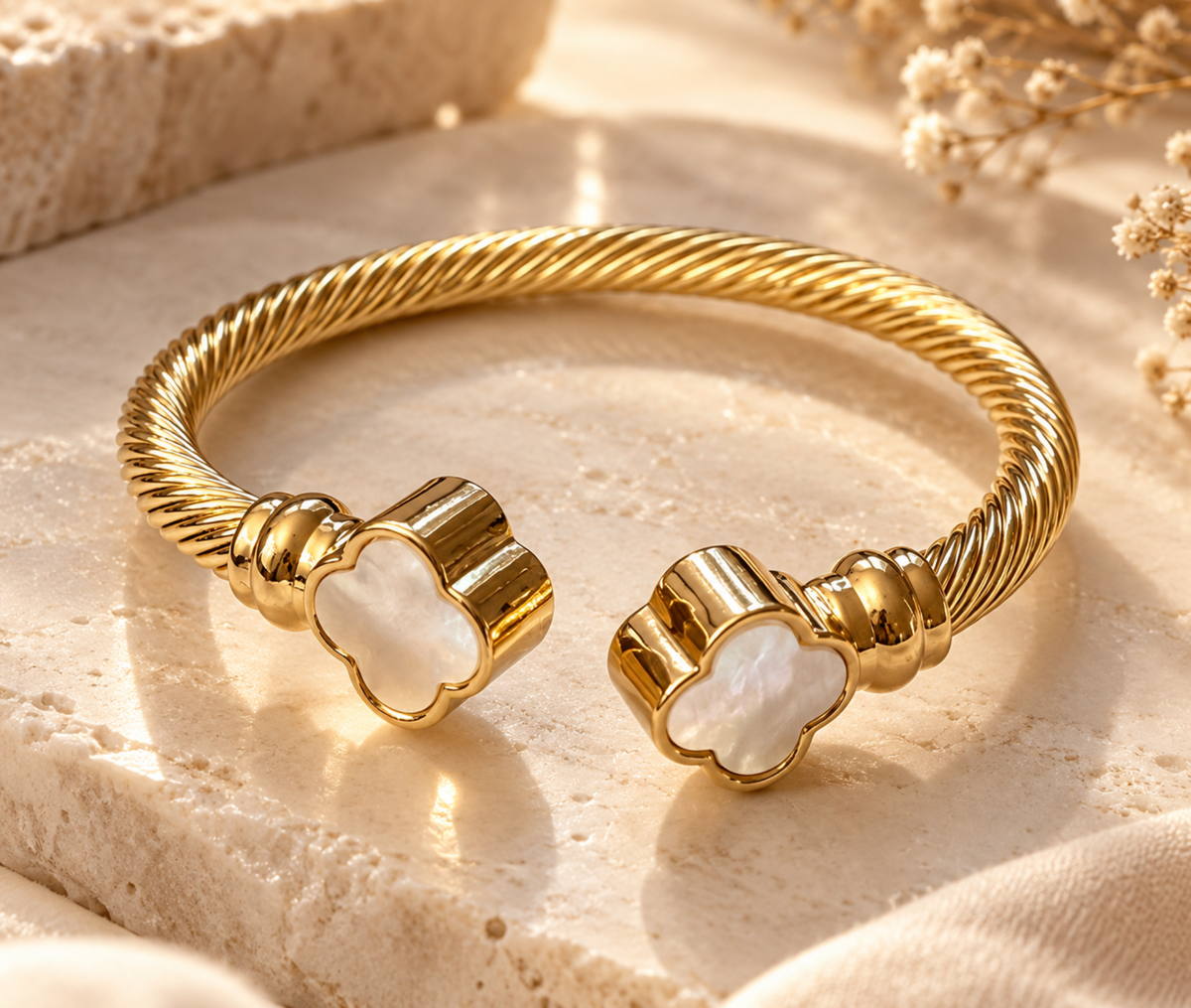 A gold-toned twisted cable cuff bracelet with two clover-shaped white mother-of-pearl end caps, displayed on a textured beige stone surface with soft dried flowers in the background.