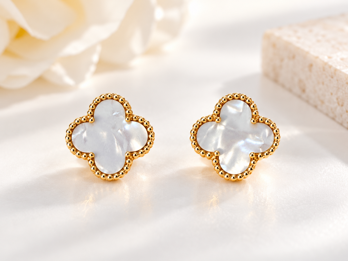 A pair of gold-tone four-leaf clover stud earrings with milky white mother-of-pearl inlays, displayed on a soft white surface with a blurred white flower and a beige stone block in the background.
