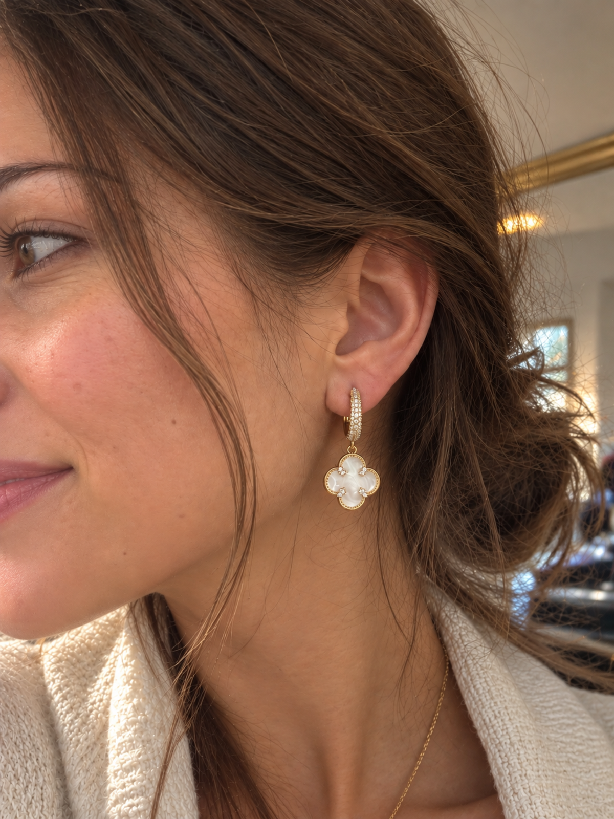 A close-up side profile of a woman wearing a gold and white clover-shaped drop earring with a small pavé band, set against an indoor background.