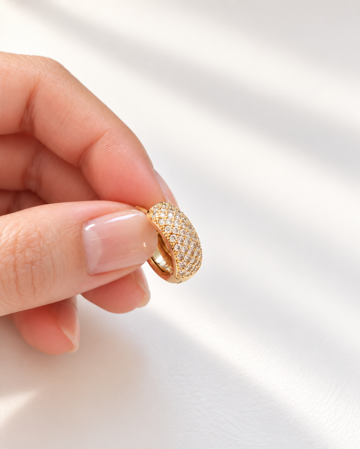 A close-up of a hand holding a gold ring paved with small clear stones against a softly lit white background.