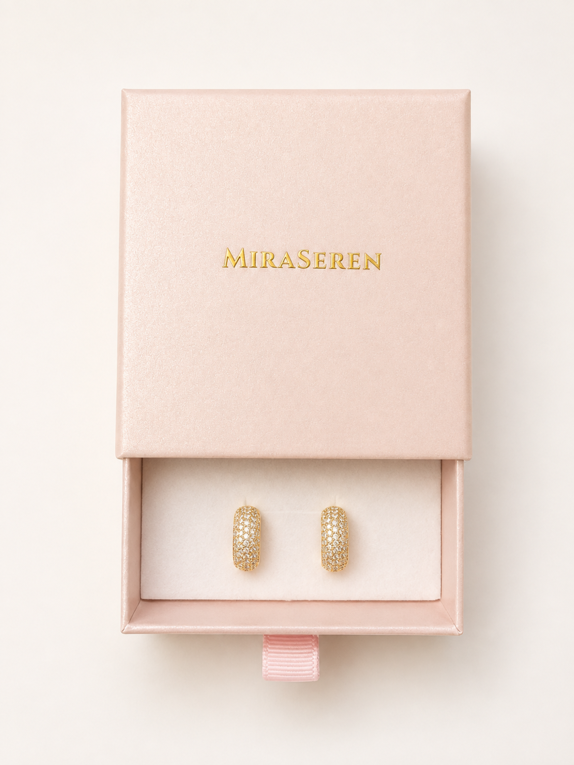 A pair of gold hoop earrings displayed inside an open blush pink Miraseren jewelry box with a ribbon pull, on a clean off-white background.