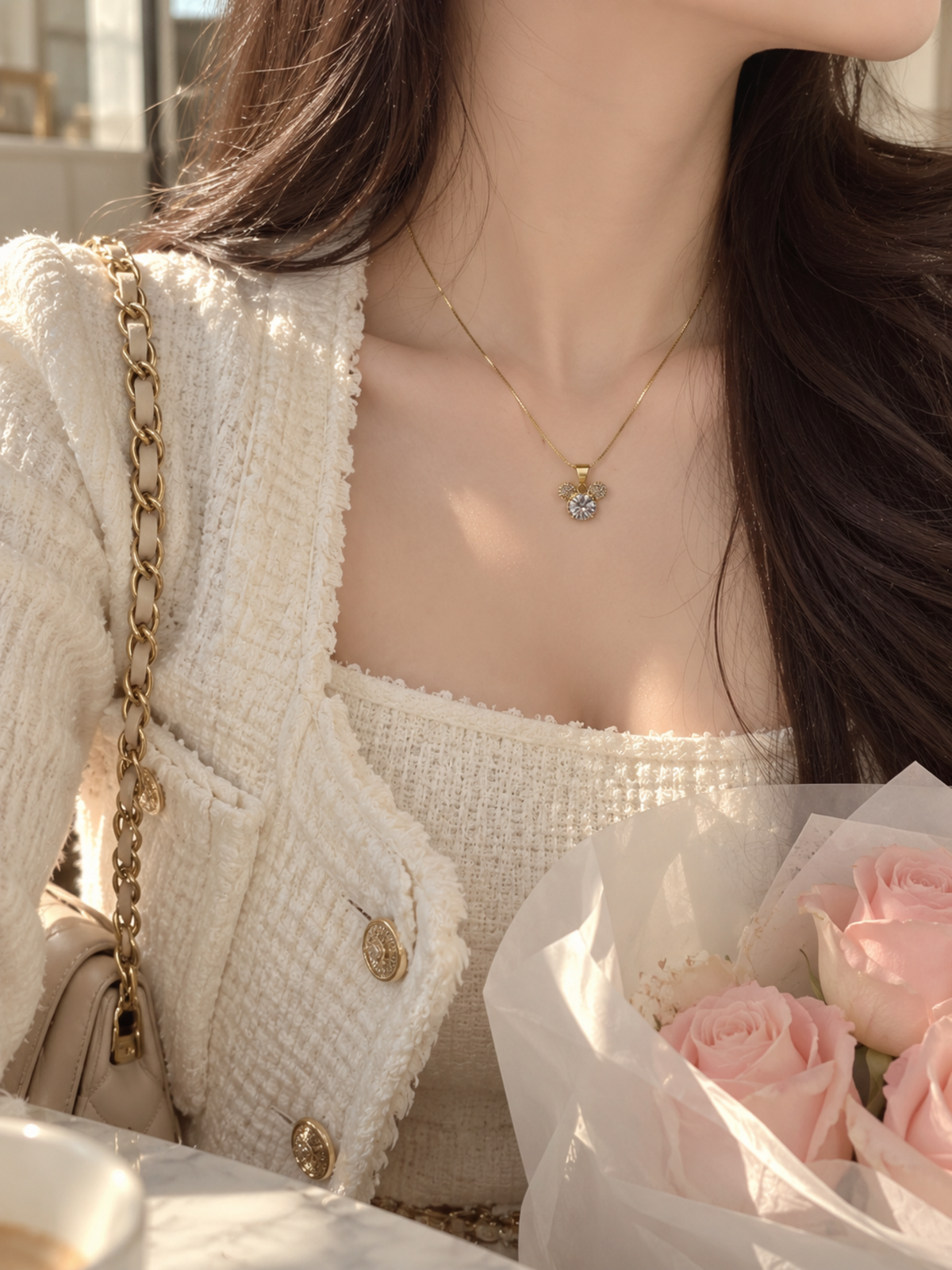 A close-up lifestyle shot of a woman wearing a delicate gold necklace with a small clear heart-shaped gemstone pendant. She is wearing a textured cream tweed jacket and top, holding a bouquet of pink roses next to a gold chain handbag.
