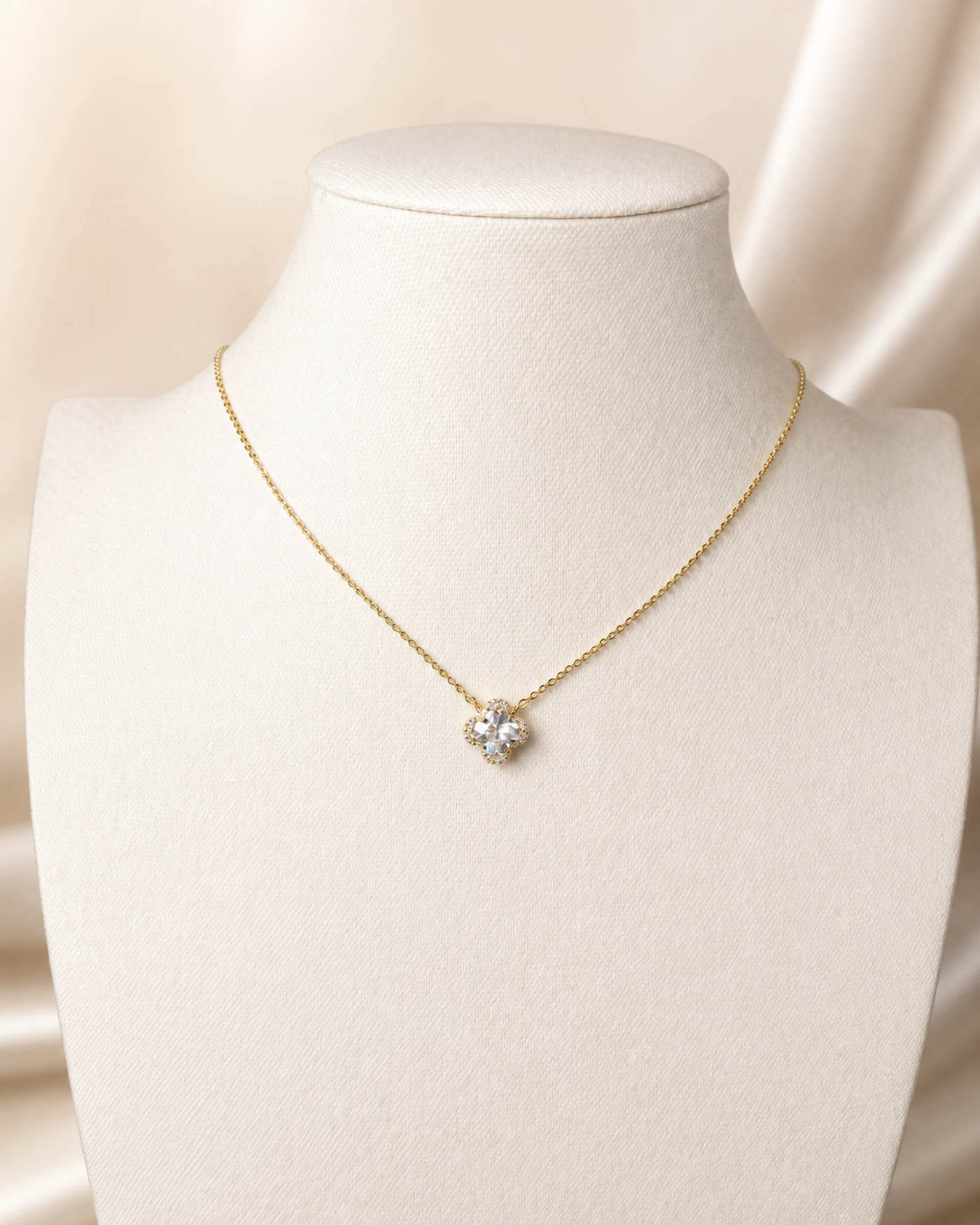 A delicate gold chain necklace with a small heart-shaped cluster of clear stones, displayed on a cream-colored jewelry bust against a soft beige satin background.