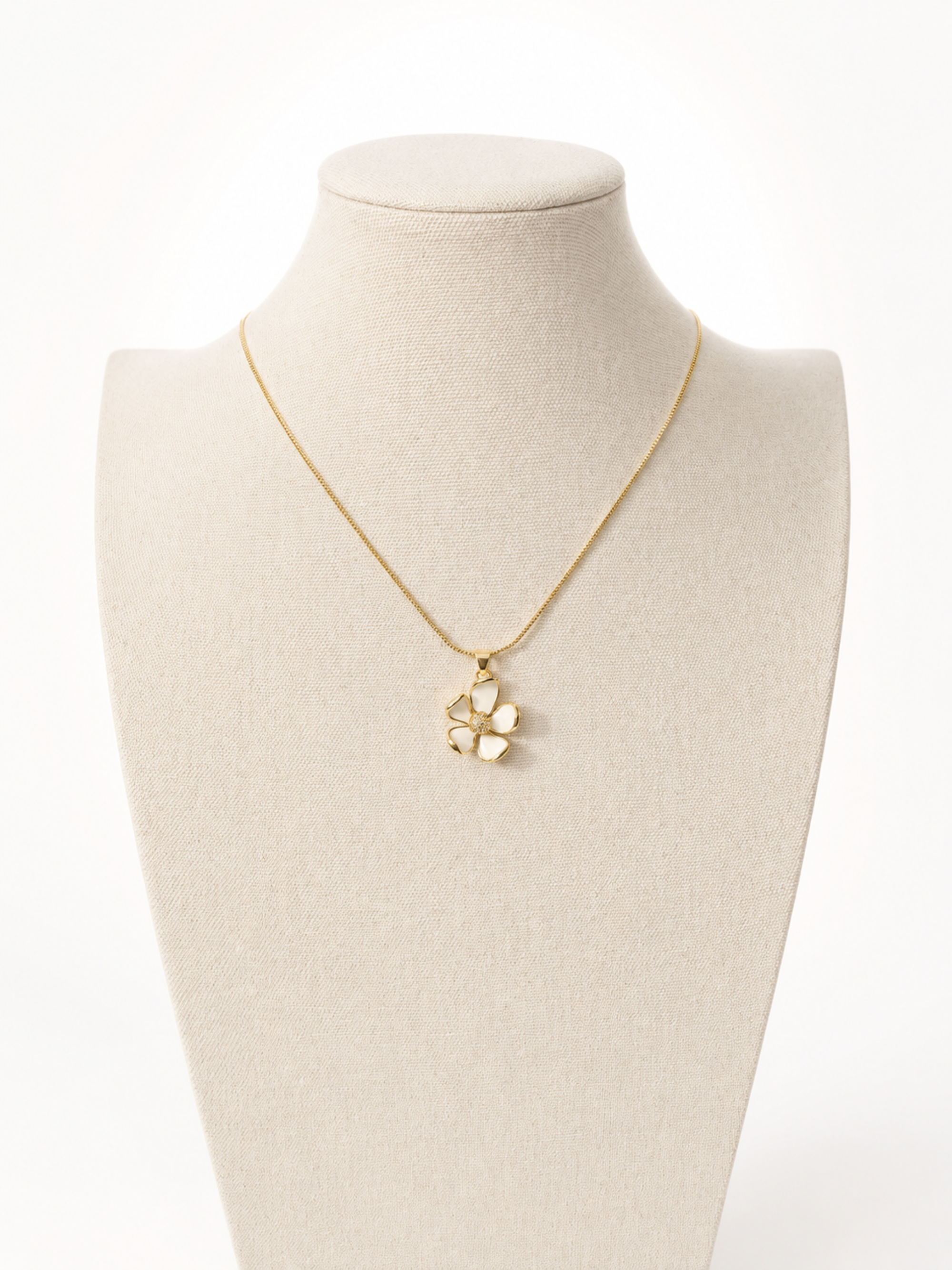 A delicate gold chain necklace with a small flower-shaped pendant, displayed on a cream-colored fabric jewelry bust against a white background.