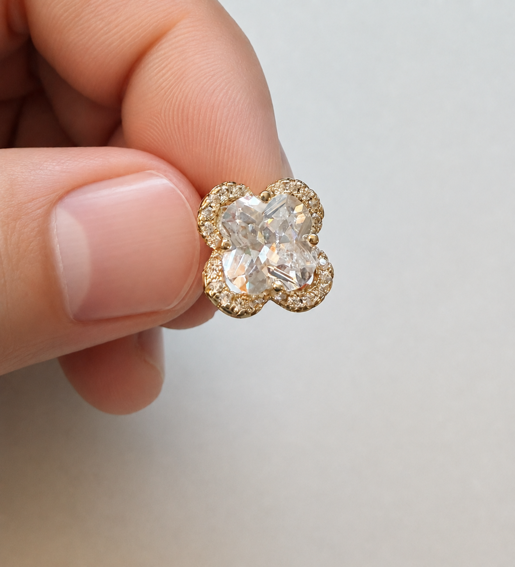 A close-up of a hand holding a small gold four-petal flower stud earring with a clear central gemstone and small clear accent stones around the edges, set against a plain light gray background.