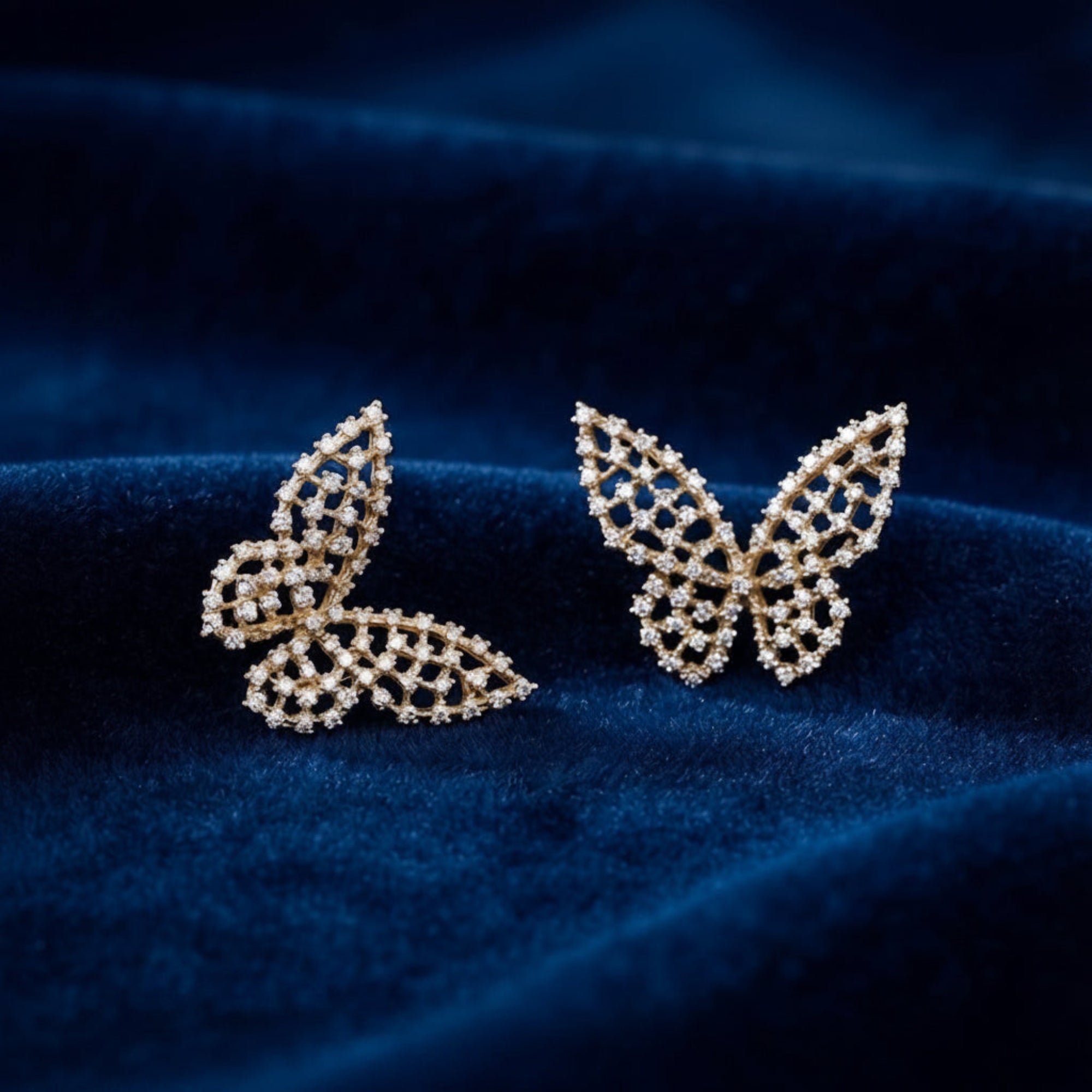A pair of butterfly-shaped stud earrings with an openwork gold-tone metal design and small clear stones, displayed on a dark navy blue velvet fabric background.