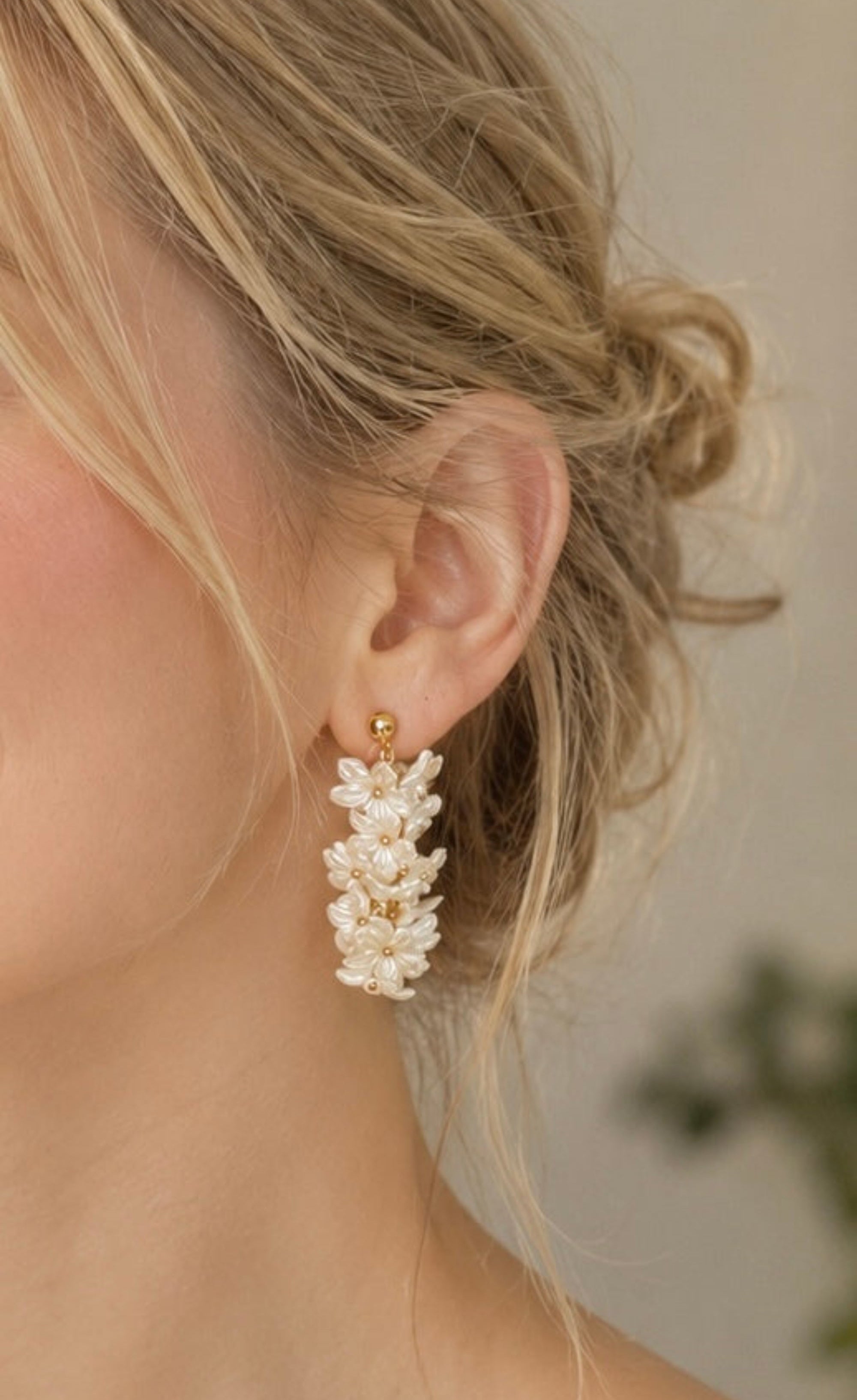 A close-up of a woman's ear wearing a dangling earring made of clustered white pearl-like beads with a small gold post, against a softly blurred neutral background.