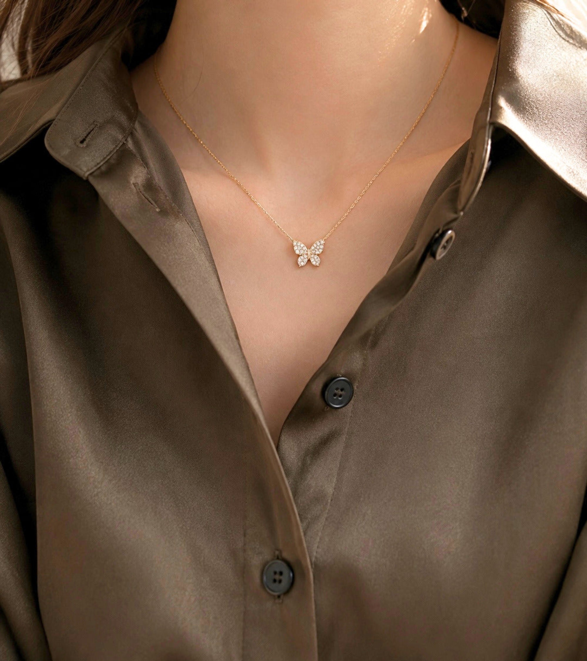 A close-up of a model wearing a delicate gold butterfly necklace with clear stones, styled with a satin brown button-down shirt.