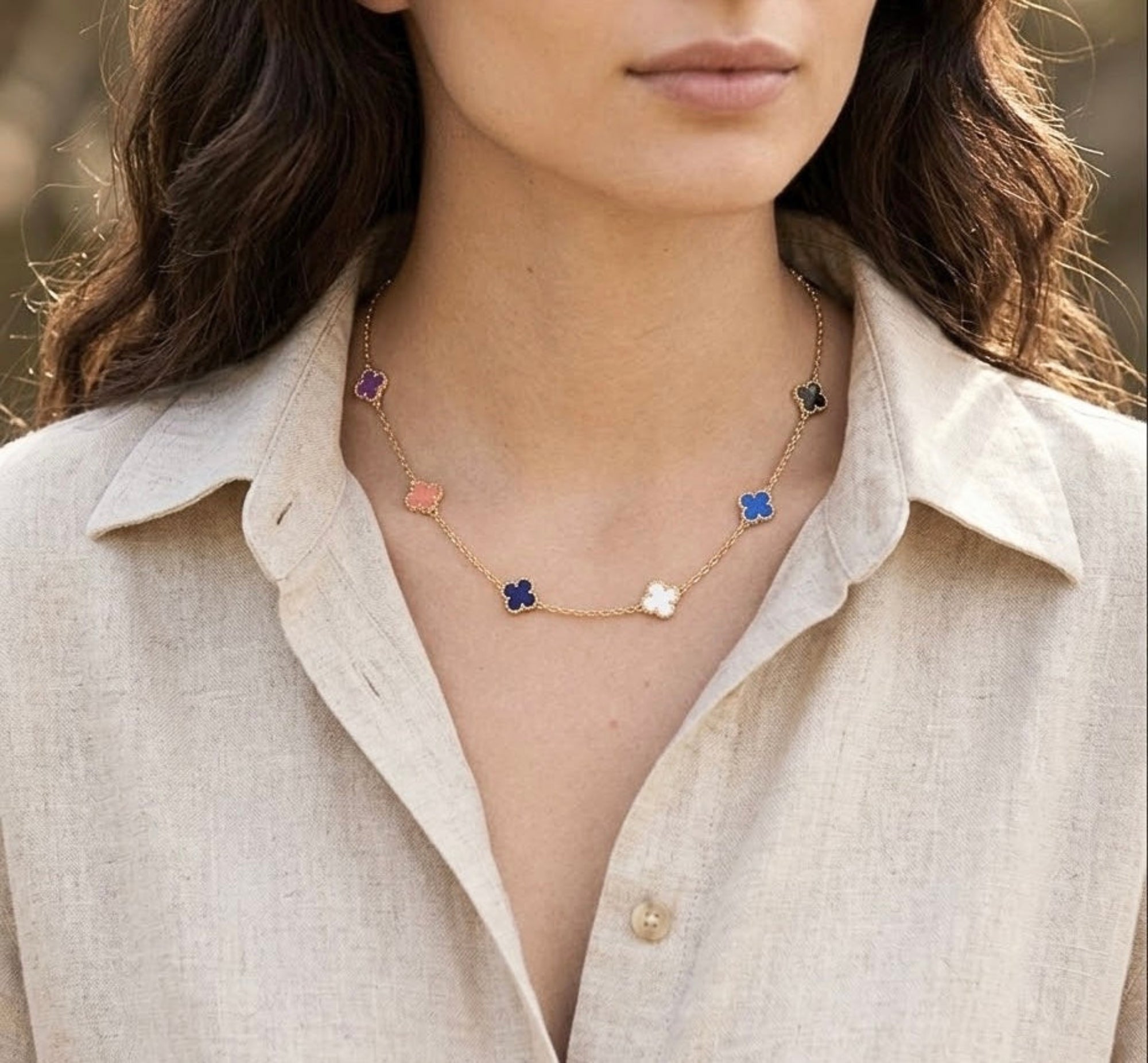 A close-up of a woman wearing a delicate gold necklace with five small colorful flower charms in purple, pink, blue, and white, styled with a beige linen shirt.