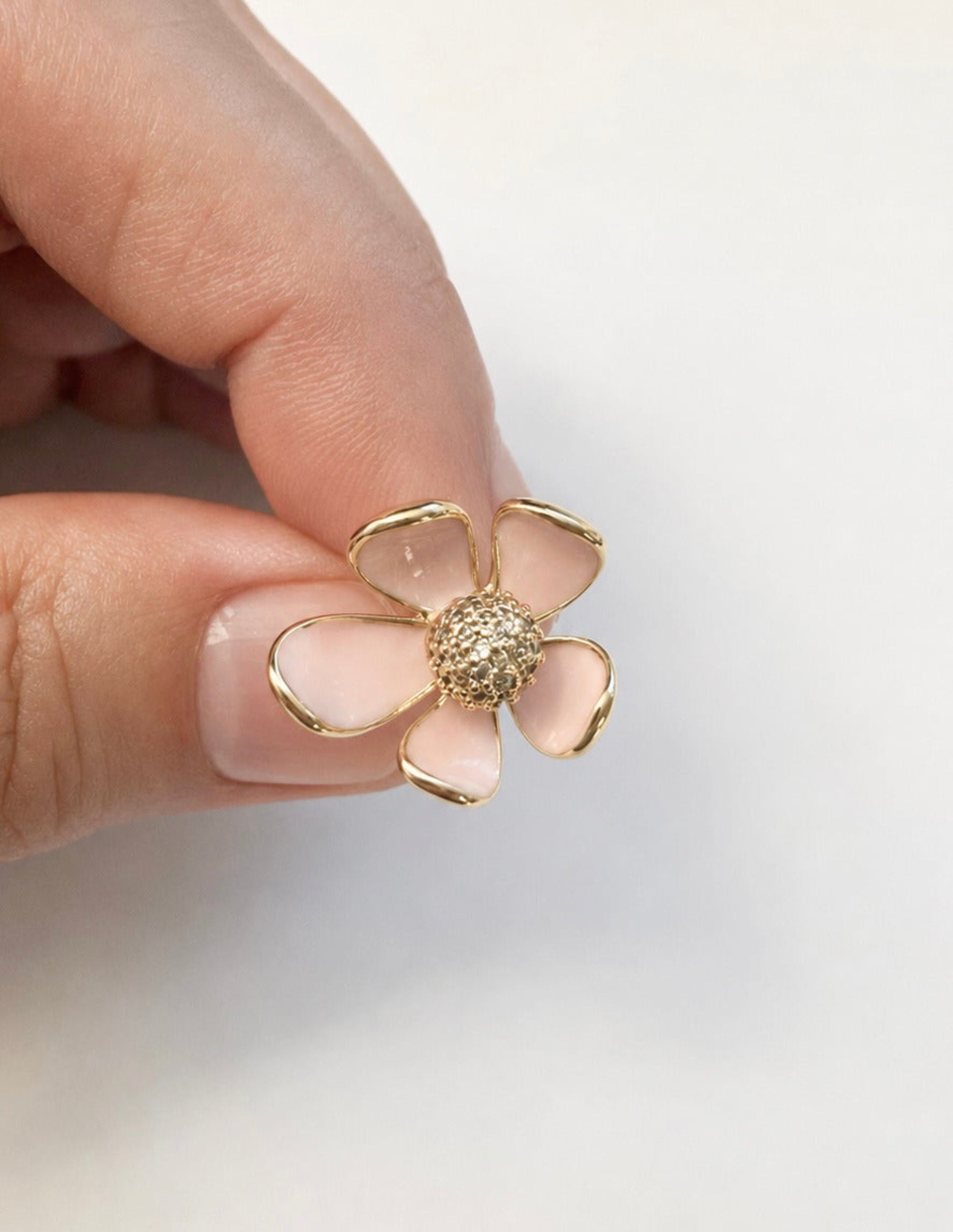 A close-up of a hand holding a small gold flower-shaped stud earring with pale pink enamel petals and a textured gold center, against a plain white background.