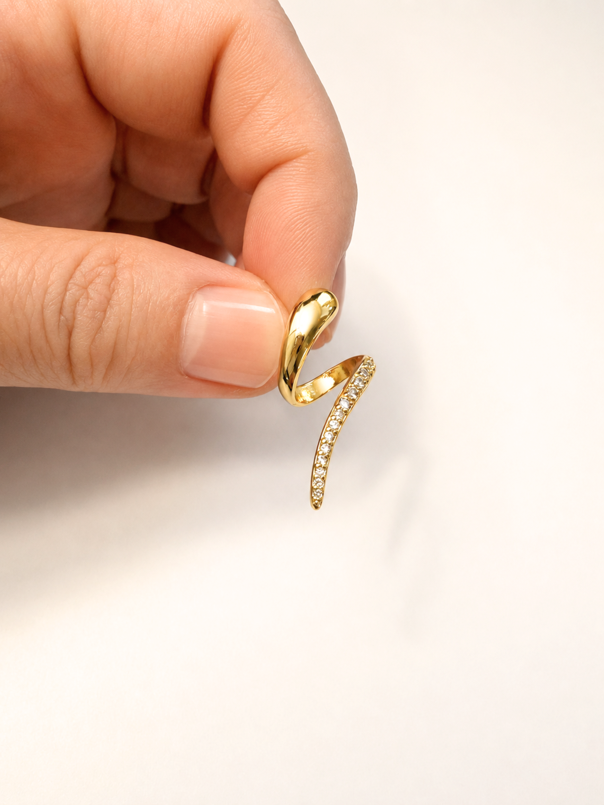 A close-up of a hand holding a gold ring with a curved, ribbon-like design set with small clear stones on a plain white background.