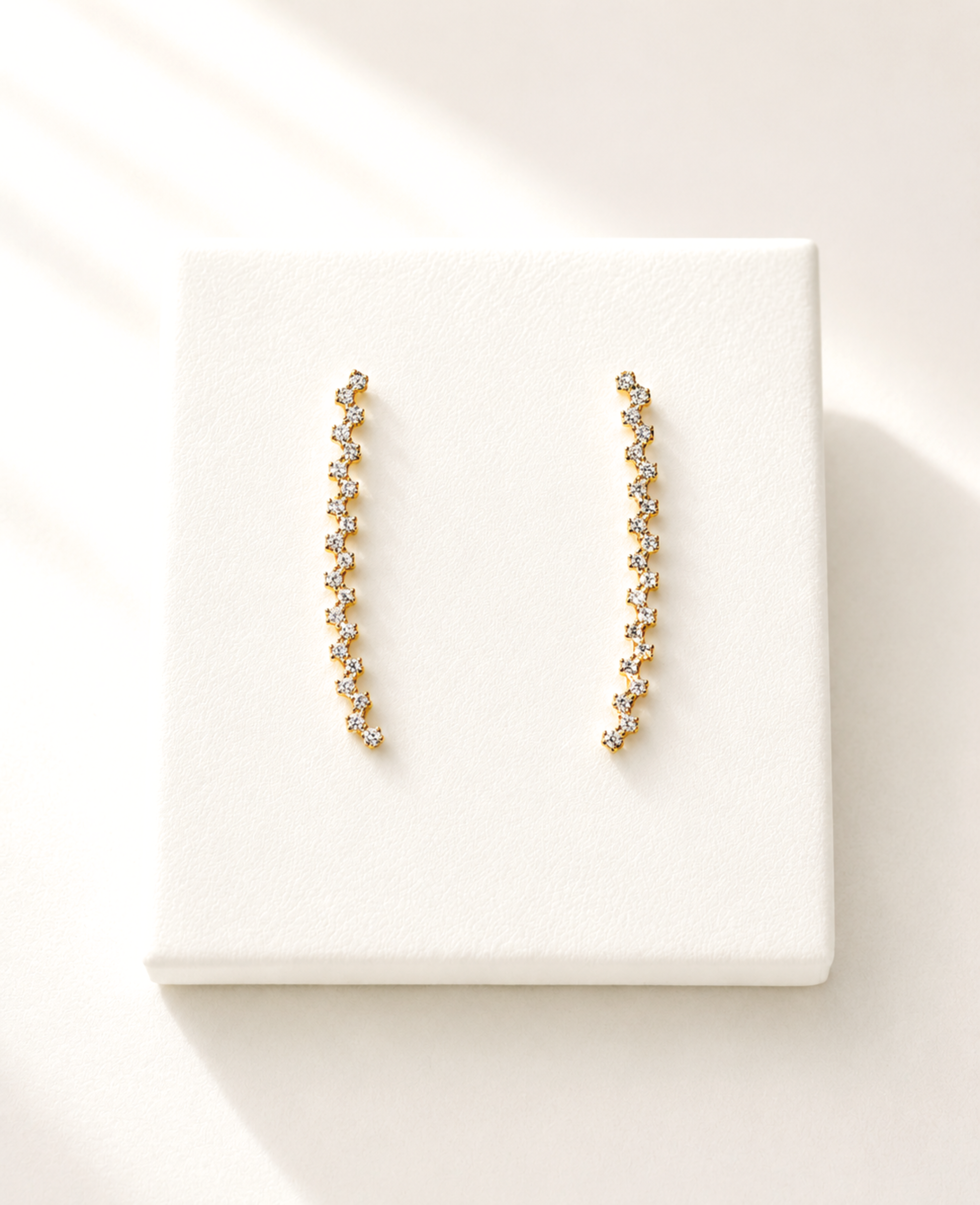 A pair of gold-toned dangle earrings with small clear stones, displayed on a white textured square card against a warm off-white background with soft shadows.