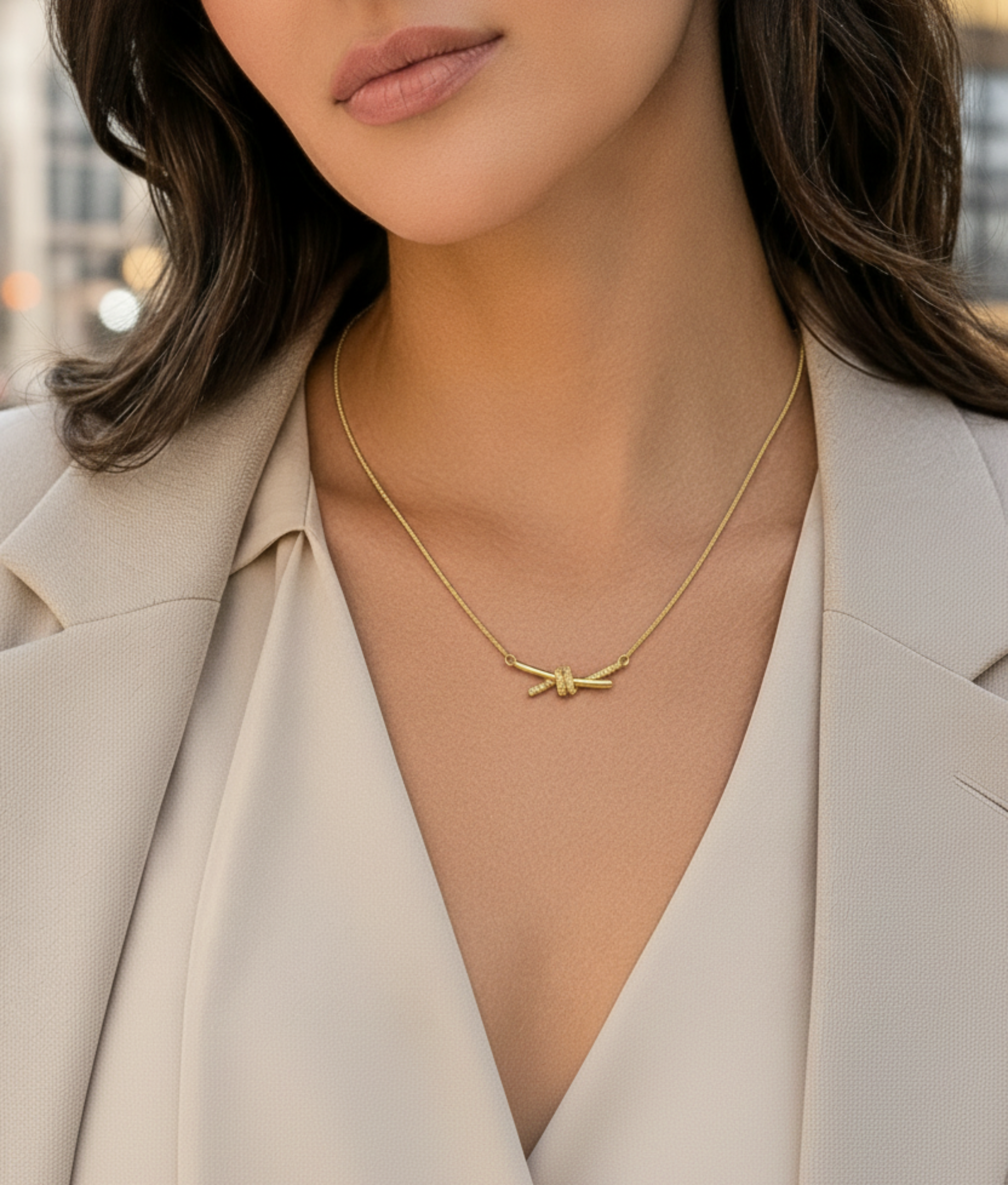 A close-up lifestyle shot of a woman wearing a delicate gold necklace with a bow pendant, styled with a beige blazer and cream blouse.