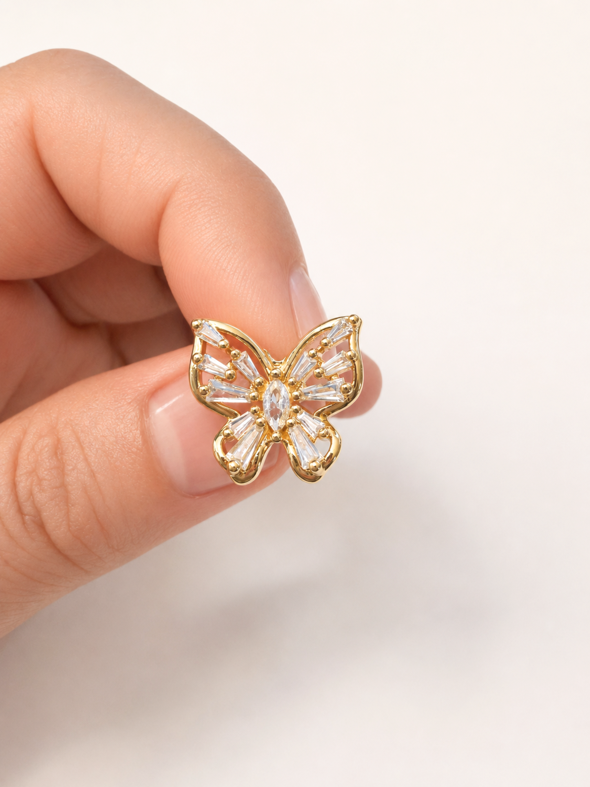 A gold butterfly-shaped stud earring with clear baguette crystals, held between a person's fingers against a plain off-white background.