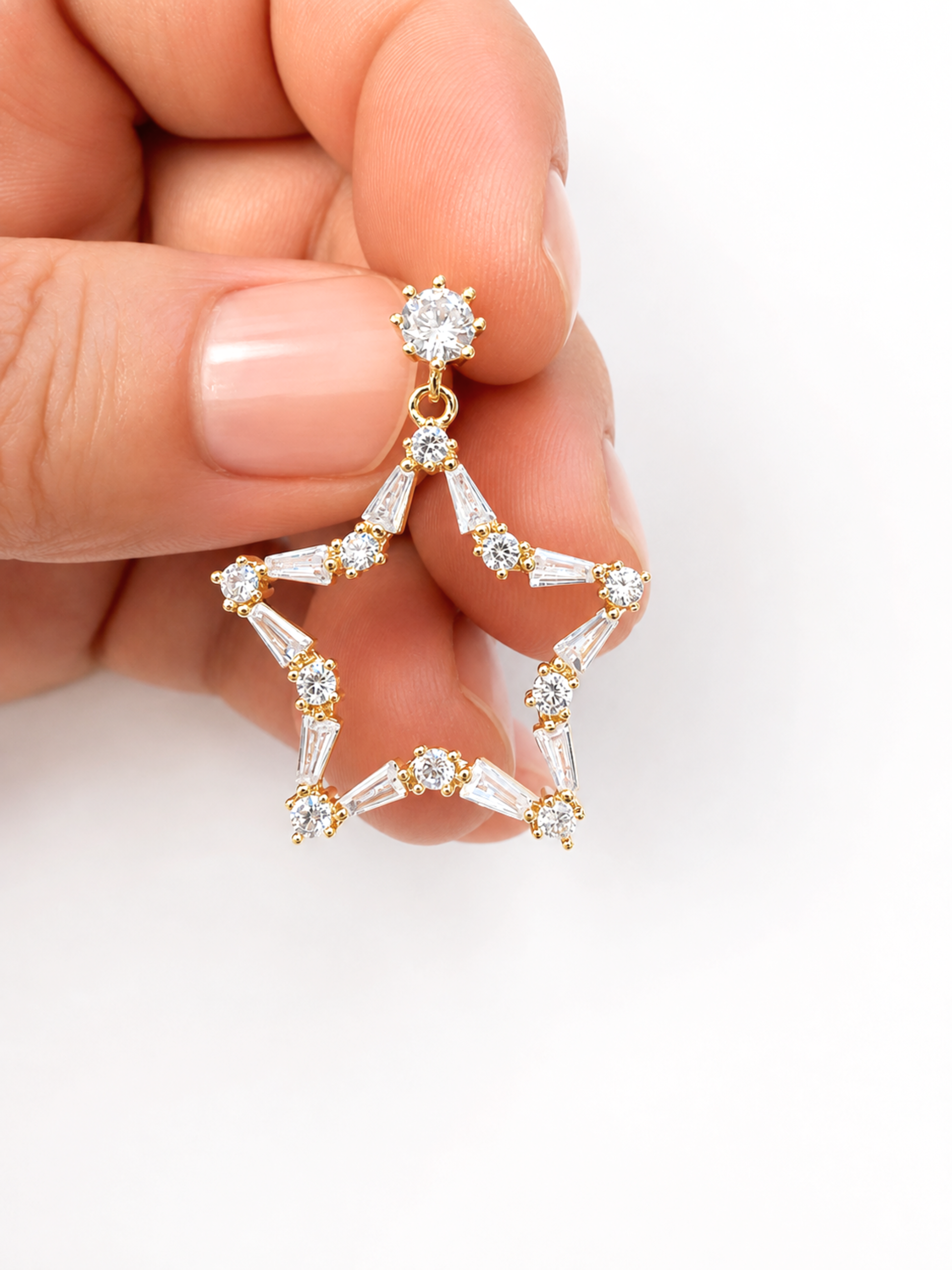 A hand with manicured nails holds a gold star-shaped dangle earring adorned with clear baguette and round-cut stones against a plain white background.