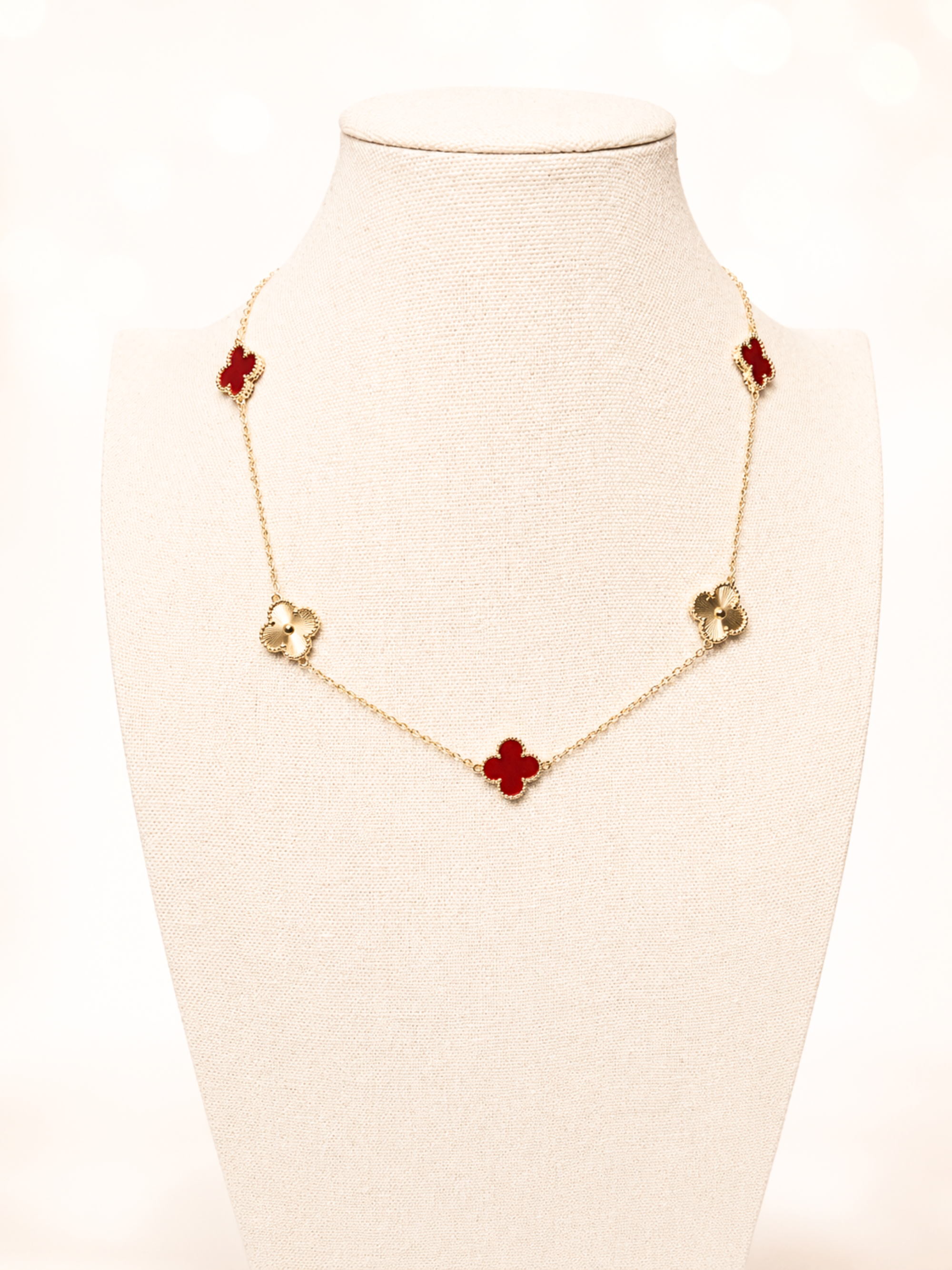 A gold-tone chain necklace with five red clover motifs, displayed on a cream-colored fabric jewelry bust against a bright, softly blurred background.