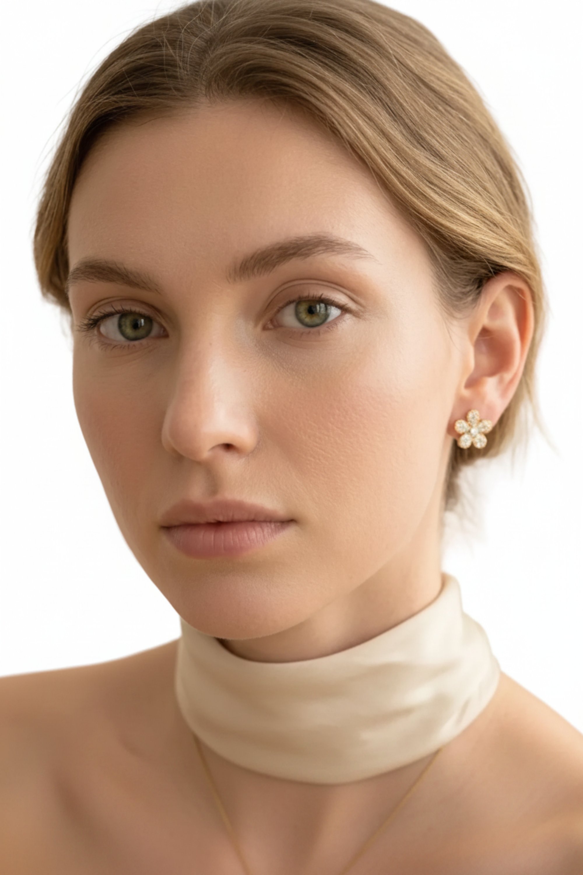 A close-up portrait of a woman with light brown hair wearing a cream satin choker and a small gold flower-shaped stud earring, set against a clean white background.