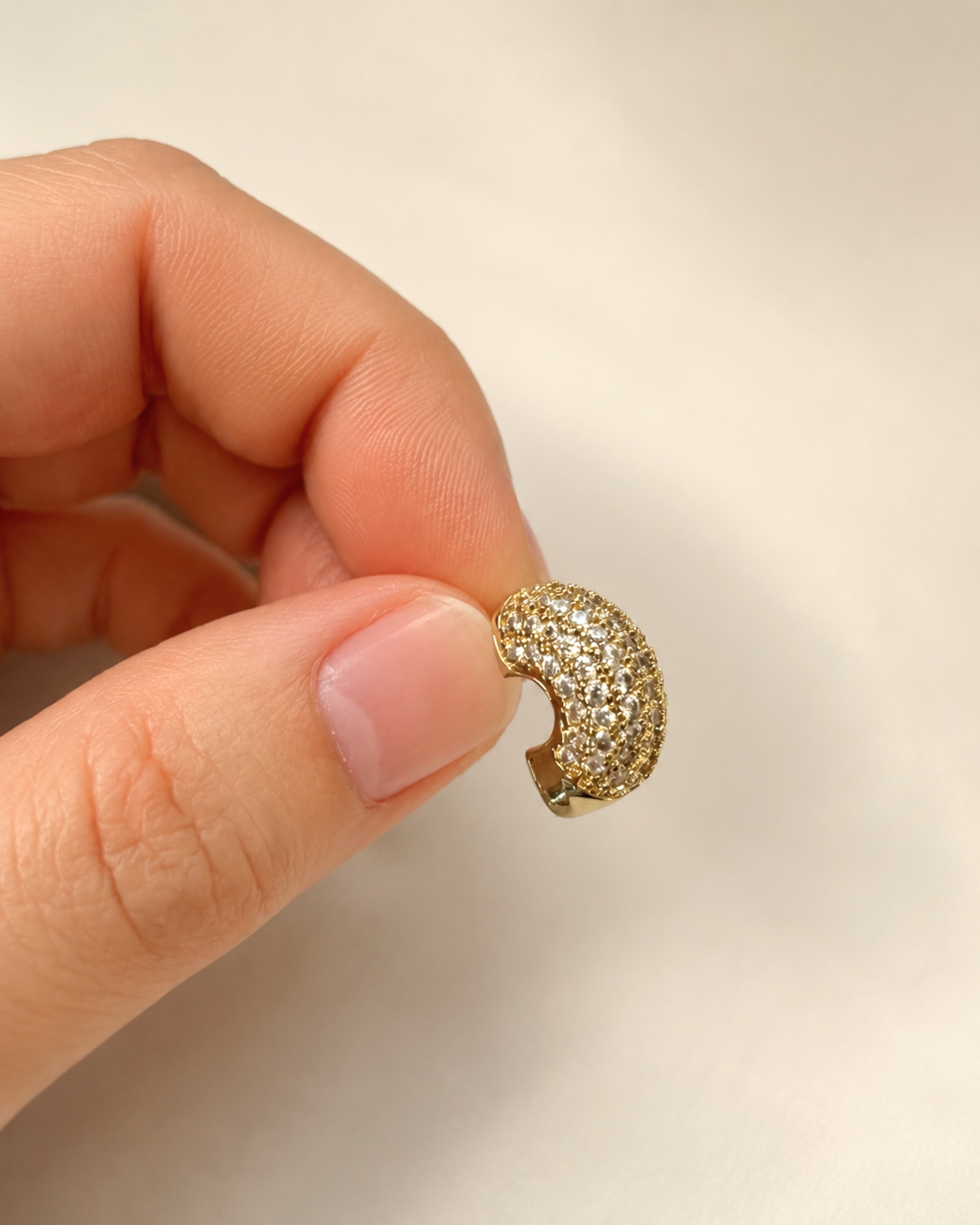 A close-up of a hand holding a small gold crescent-shaped earring paved with clear stones against a soft beige background.