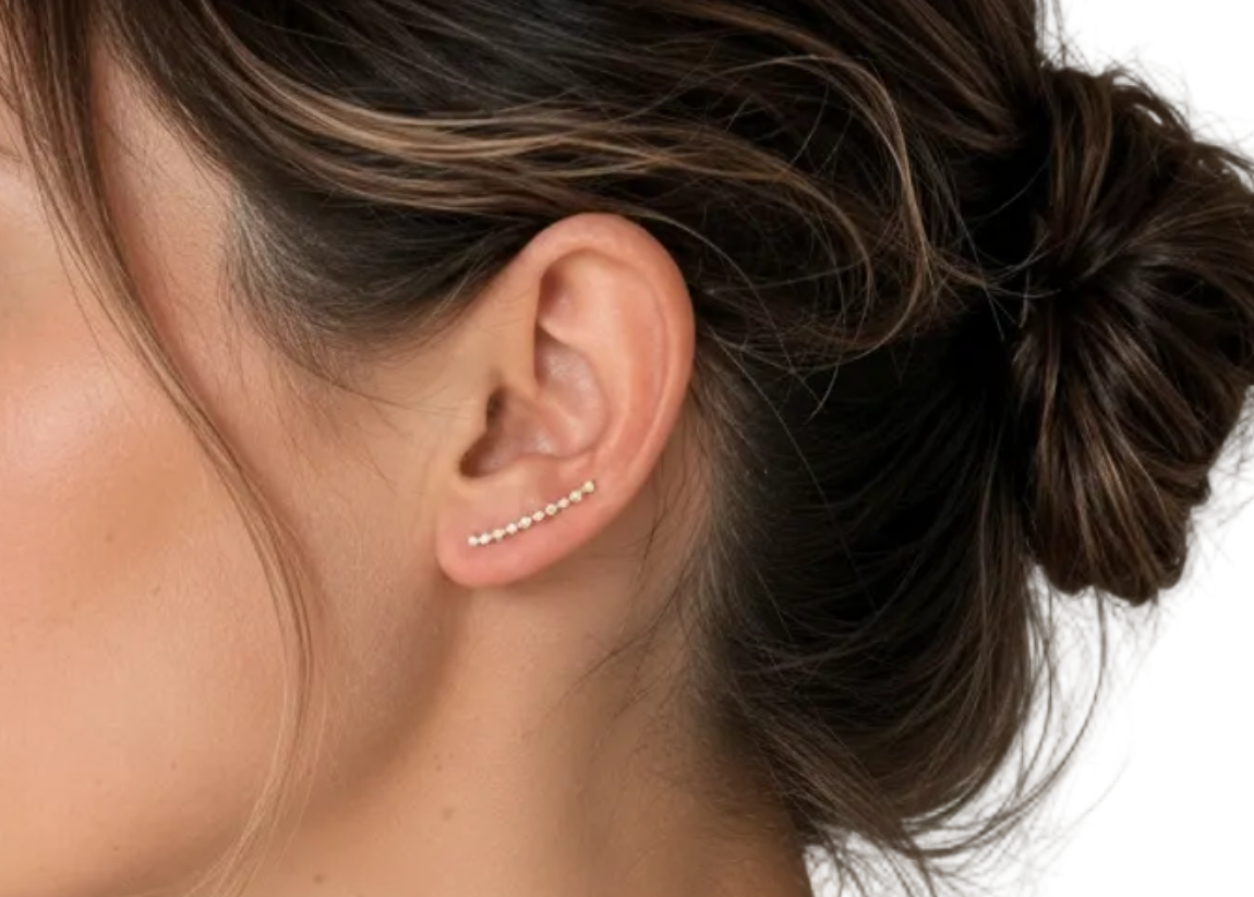 A woman in profile wearing a delicate curved ear cuff with small clear stones on her earlobe, shown against a soft neutral indoor background.