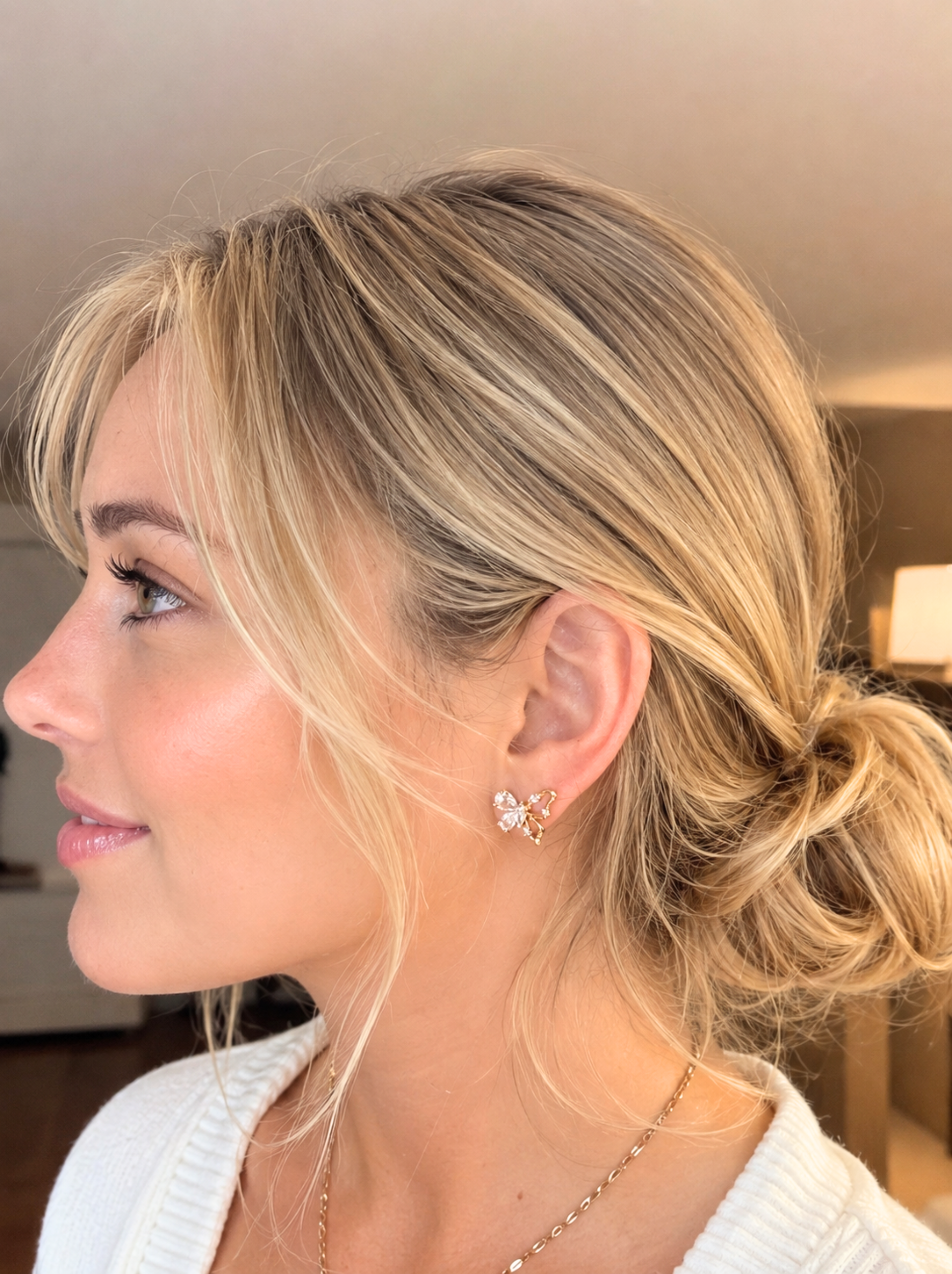 A close-up side profile of a woman with blonde hair in a low bun, wearing a small silver butterfly stud earring and a delicate gold chain necklace.