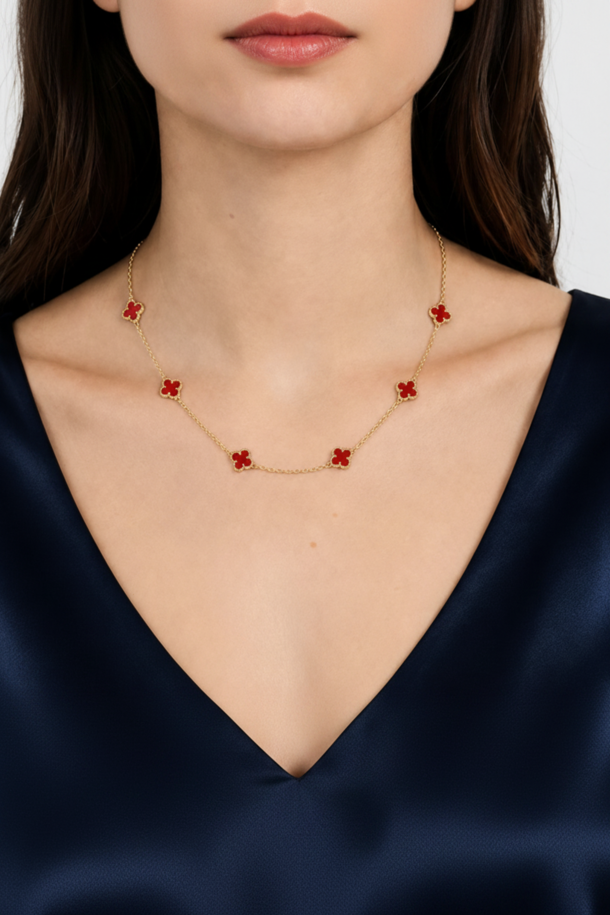 A close-up of a model wearing a delicate gold chain necklace with five small red flower-shaped charms, paired with a dark navy satin V-neck top.
