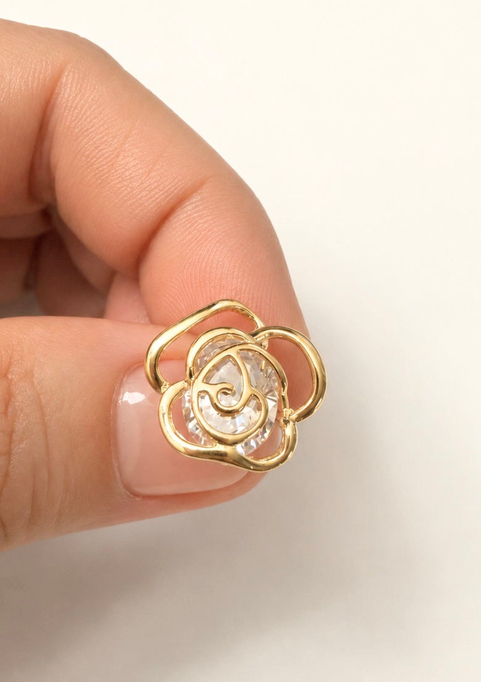 A close-up of a hand holding a small gold flower-shaped stud earring with a clear textured center, against a plain off-white background.